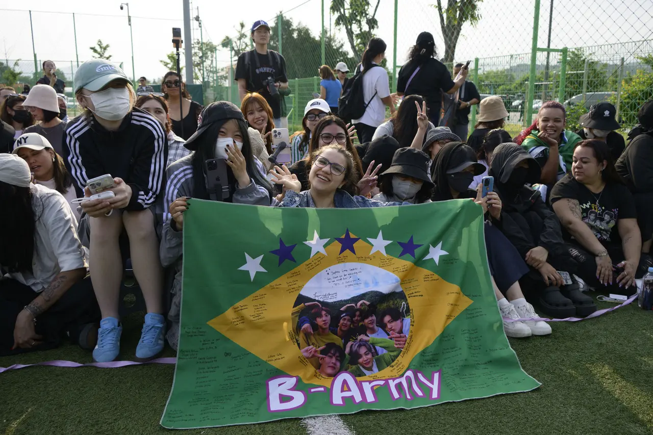 Imagem mostra grupo de fãs do BTS, fandom conhecido como ARMY, aguardando dispensa de RM e V do Exército. Uma delas segura uma bandeira brasileira com o escrito B-army.