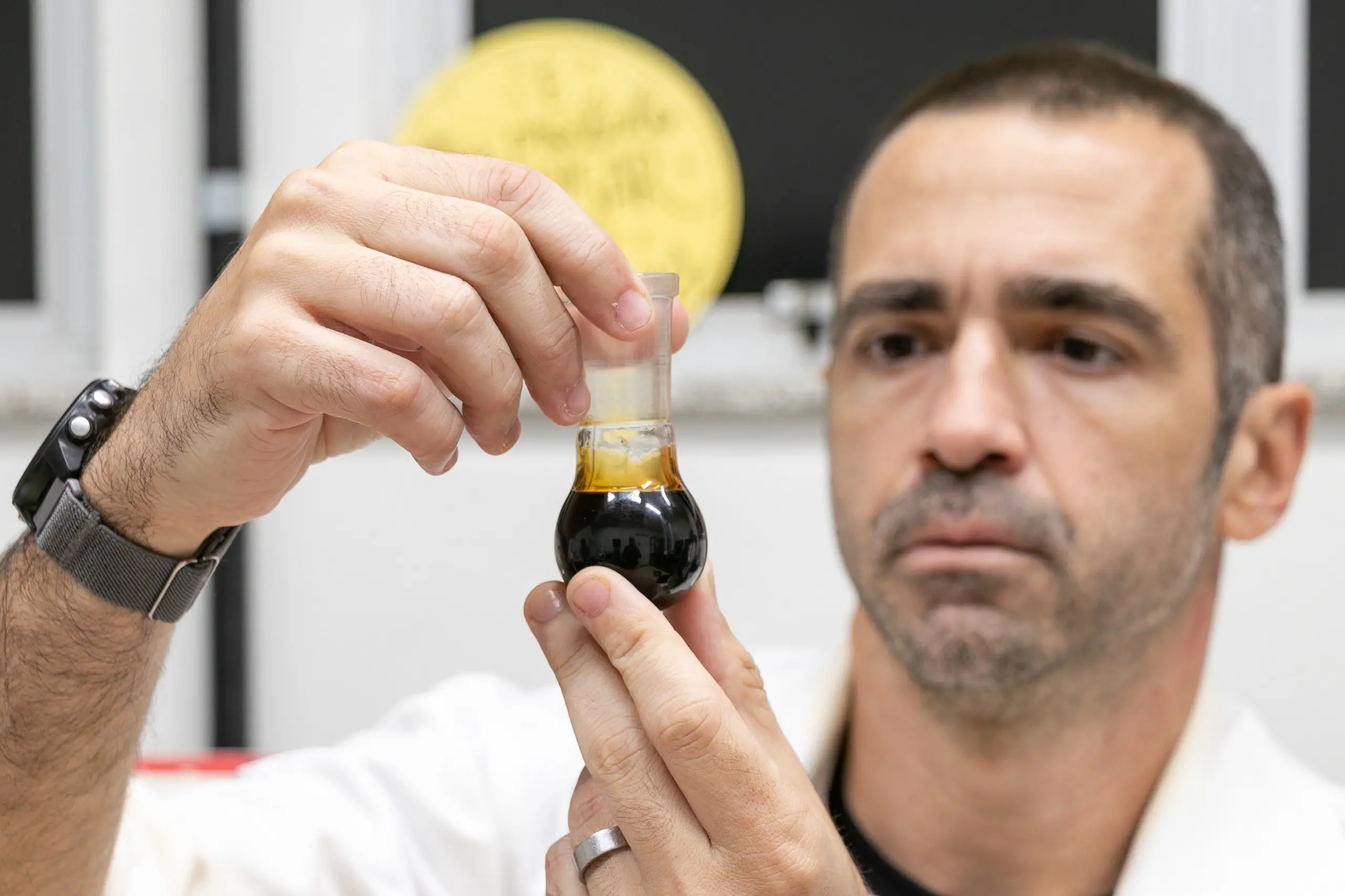 A imagem mostra um homem cientista, usando um jaleco branco. Ele segura com as duas mãos um pequeno recipiente de vidro em formato de gota, que contém um líquido de duas fases: uma camada superior amarelada e uma camada inferior escura, quase preta. O homem tem cabelos curtos e escuros, barba por fazer e uma expressão concentrada enquanto observa o conteúdo do recipiente. Ao fundo, parcialmente desfocado, pode-se ver uma superfície amarela e uma área branca que lembra uma parede ou equipamento de laboratório.