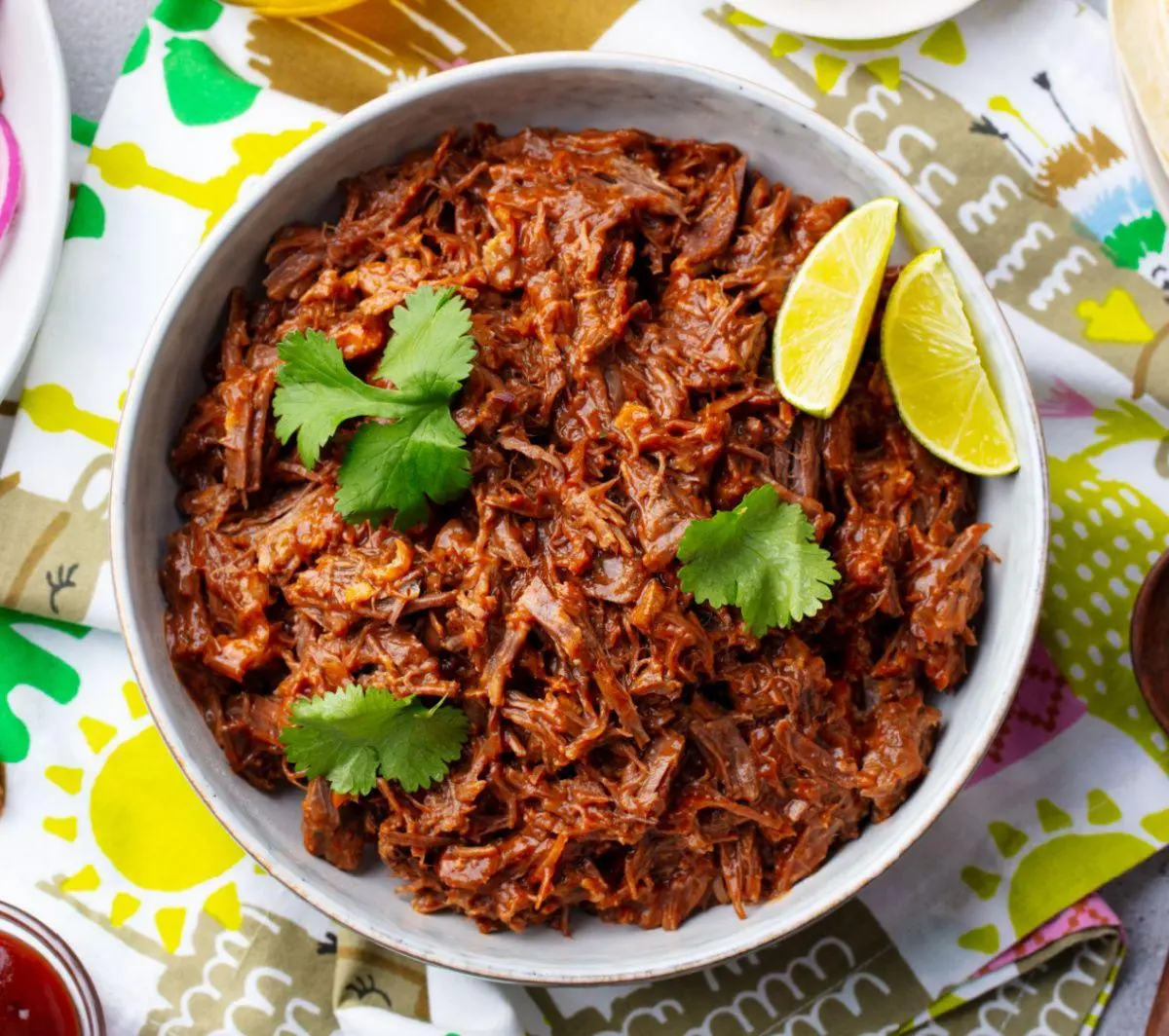Carne desfiada com limão em uma tigela branca, decorada com folhas de coentro, sobre uma toalha colorida. Prato típico brasileiro, ideal para refeições em família.