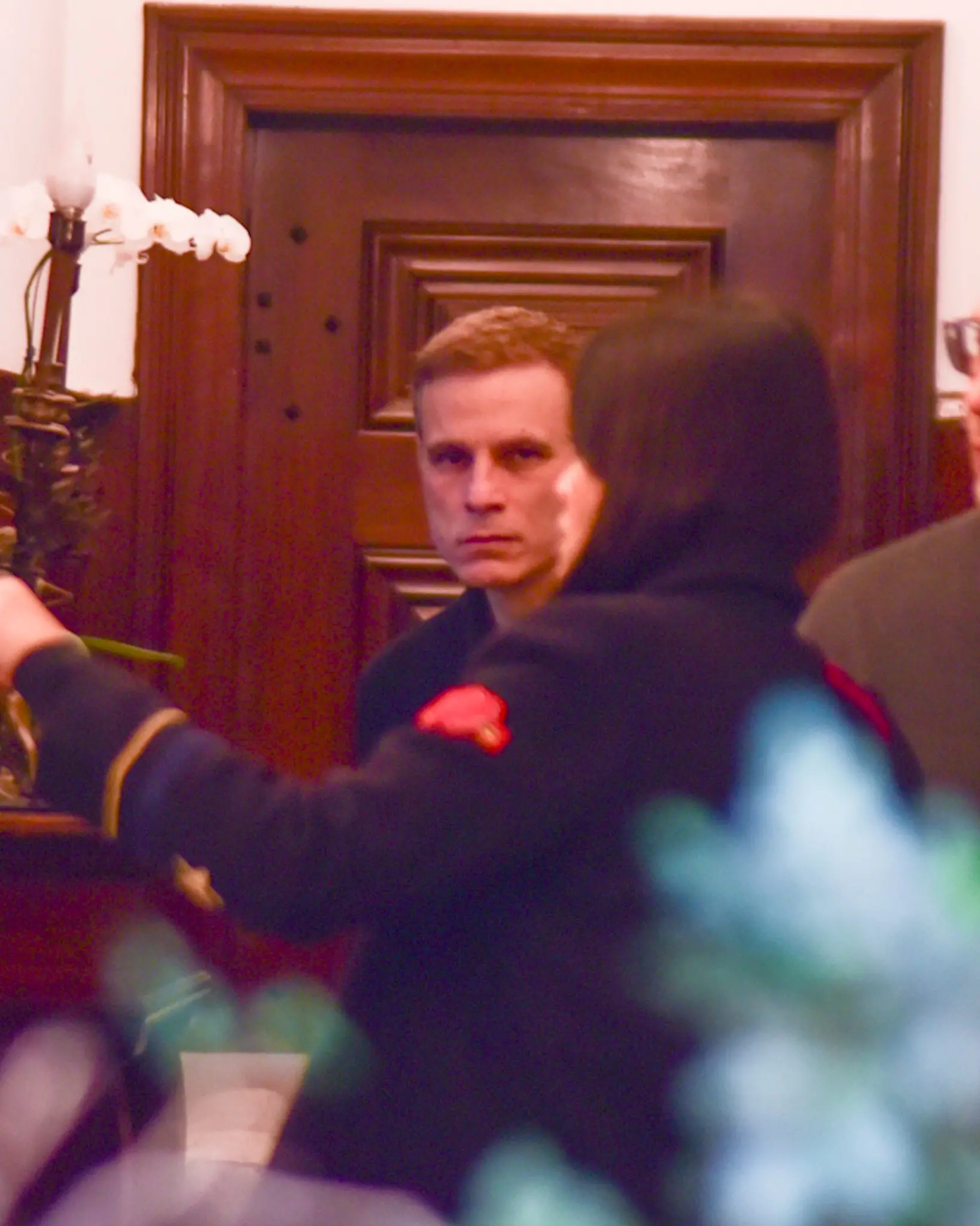 Cena do filho de Francisco Cuoco com expressão séria em um ambiente interno, com uma porta de madeira ao fundo, mostrando um momento de atenção ou curiosidade.