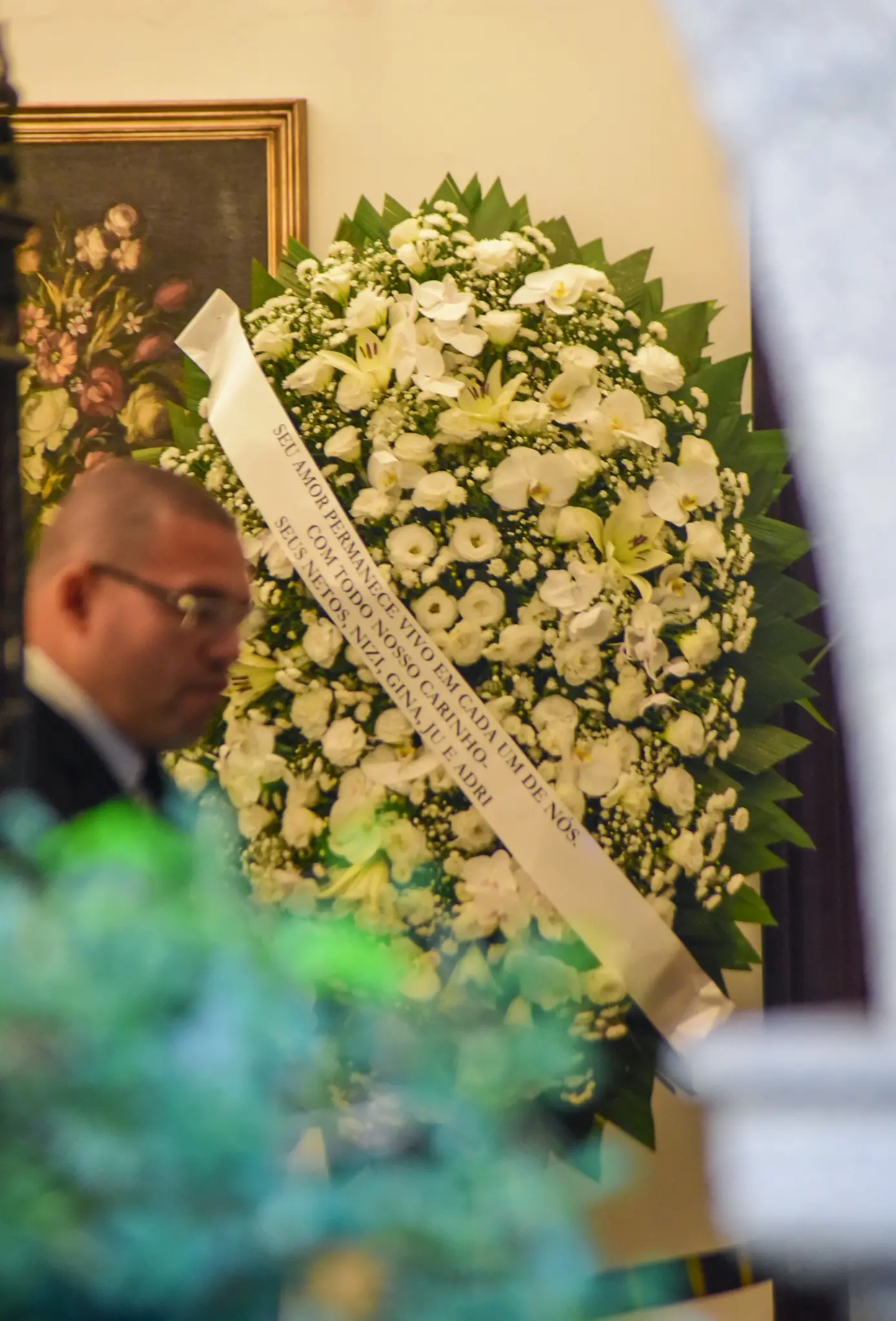 Homenagem com coroa de flores brancas em funeral de Francisco Cuoco, símbolo de condolências e respeito, com pessoa presente na cerimônia, refletindo homenagem e despedida.