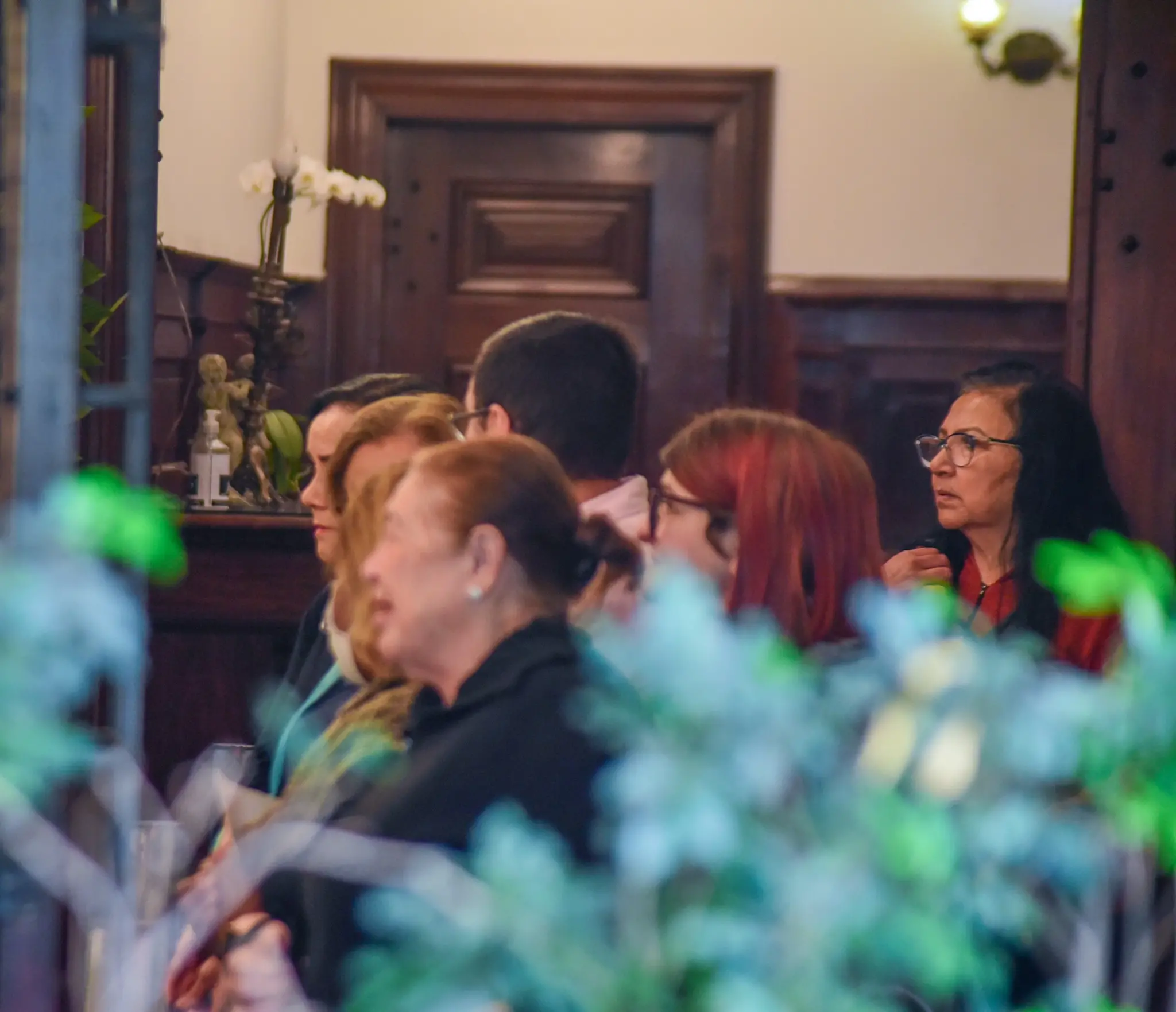 Reunião de pessoas concentradas em evento indoor, com decoração em madeira, flores e iluminação suave, sugerindo uma atmosfera de conferência ou palestra.
