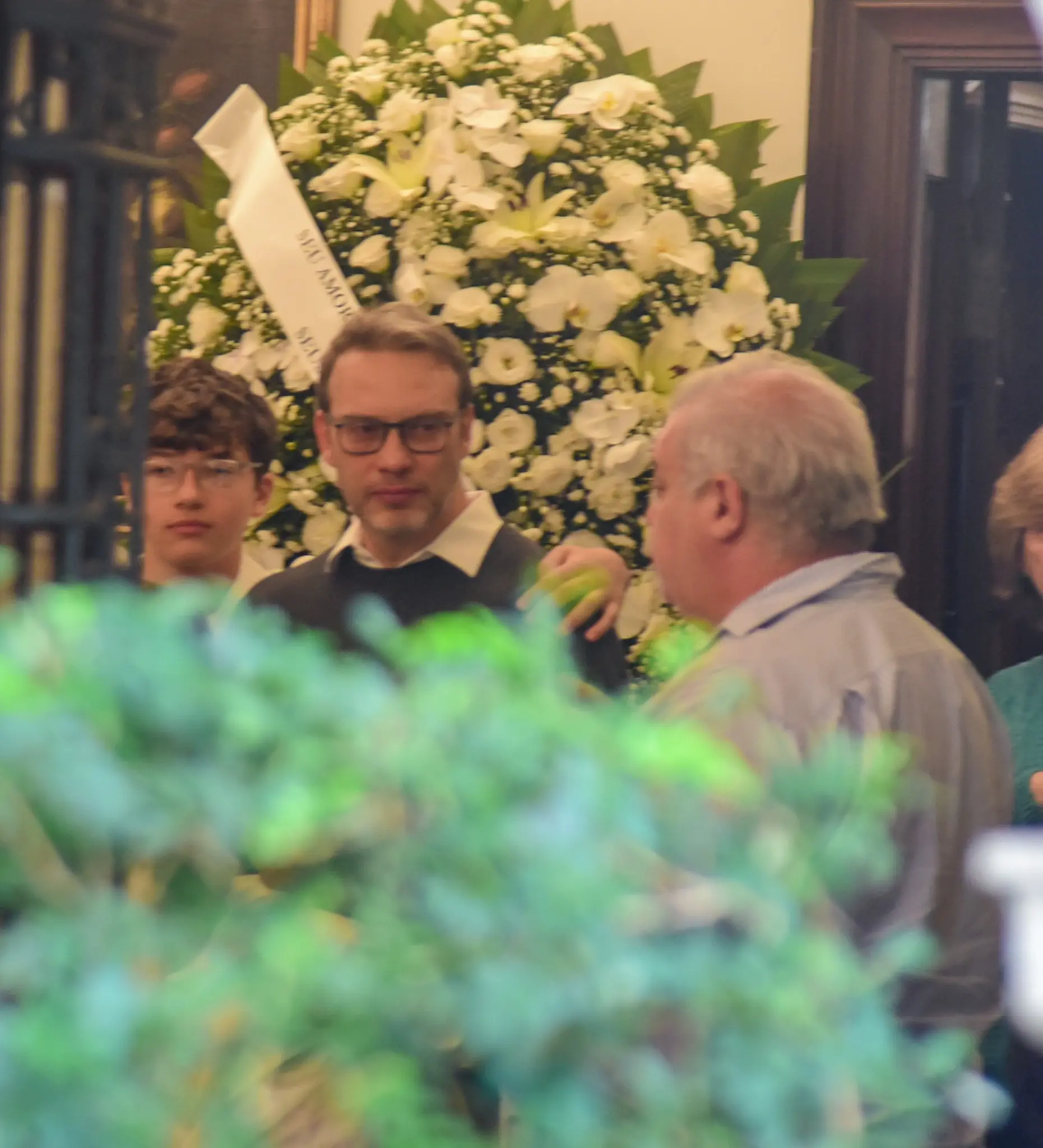Homenagem com flores brancas no funeral de Francisco Cuoco com várias pessoas conversando em um ambiente interno