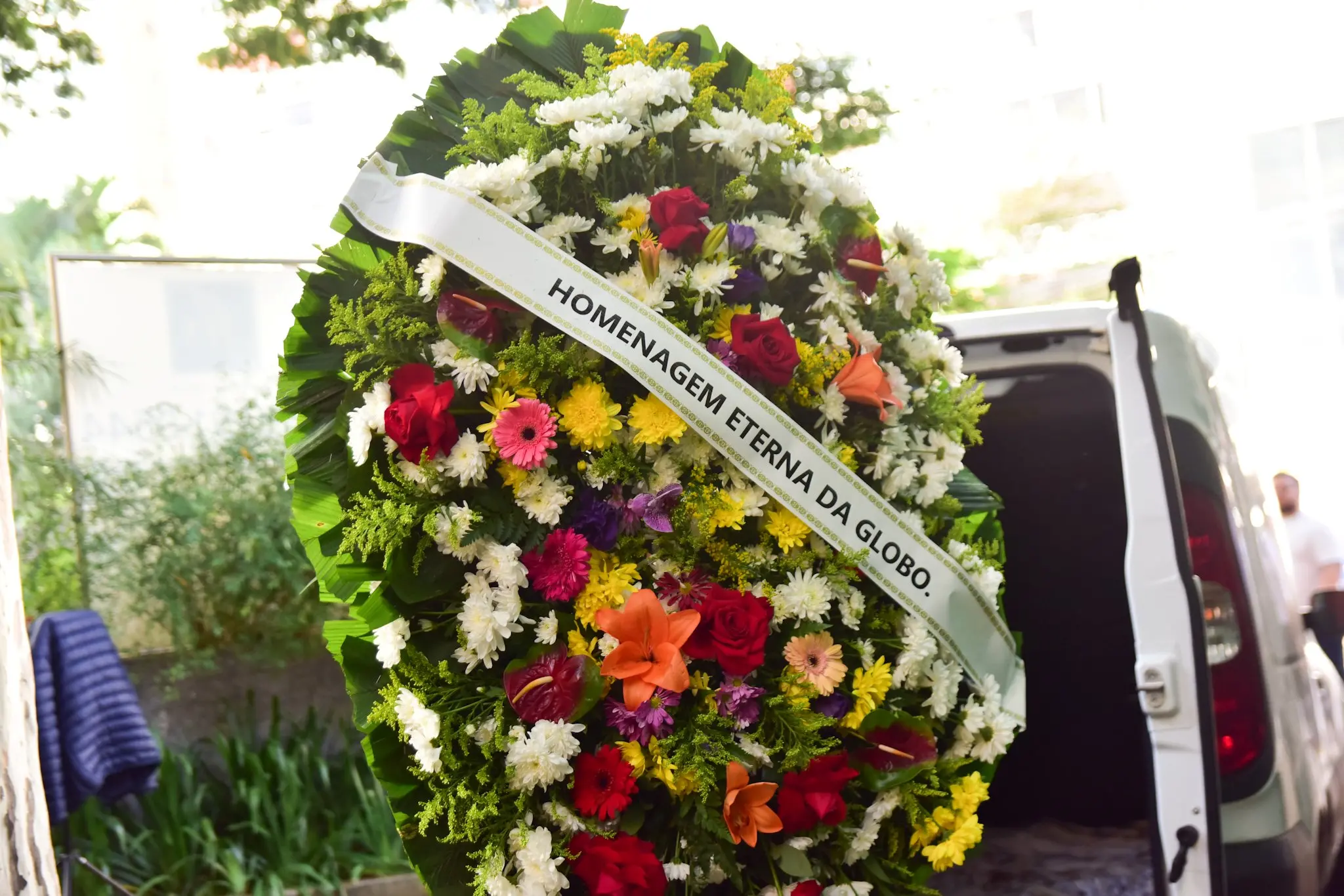 Arranjo de flores em homenagem eterna da Globo, com cores vibrantes de rosas vermelhas, orquídeas laranjas, margaridas brancas, e outras flores, em um ambiente ao ar livre, homenageando alguém especial.