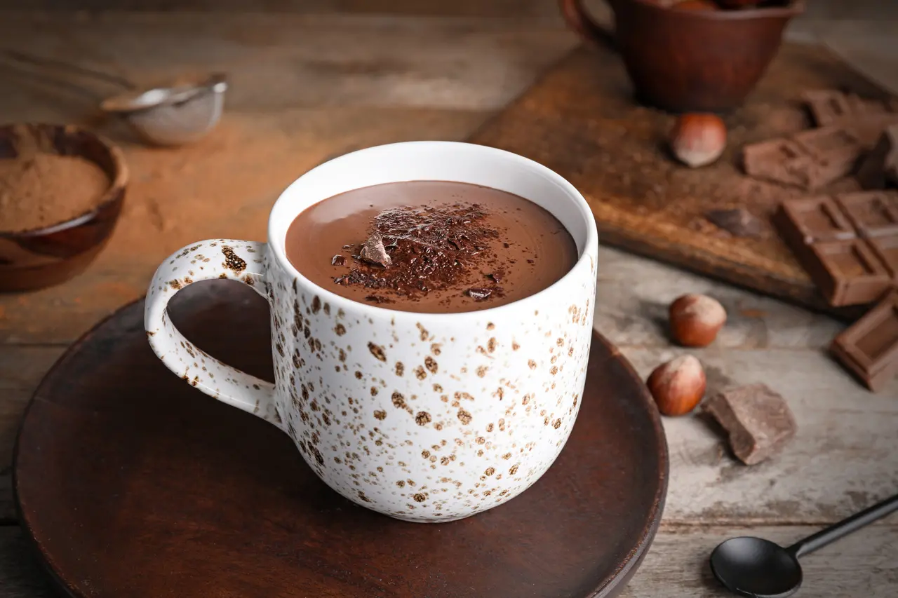 Imagem mostra xícara de chocolate quente sobre mesa de madeira, ilustrando receitas para esquentar frio no inverno 2025