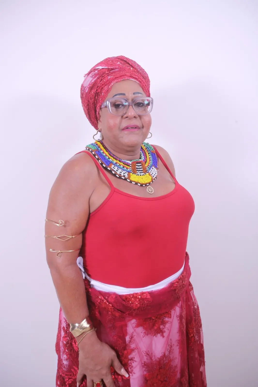 Viviane de Oya, mulher negra com turbante vermelho, colares coloridos e roupas tradicionais posa em frente a fundo branco