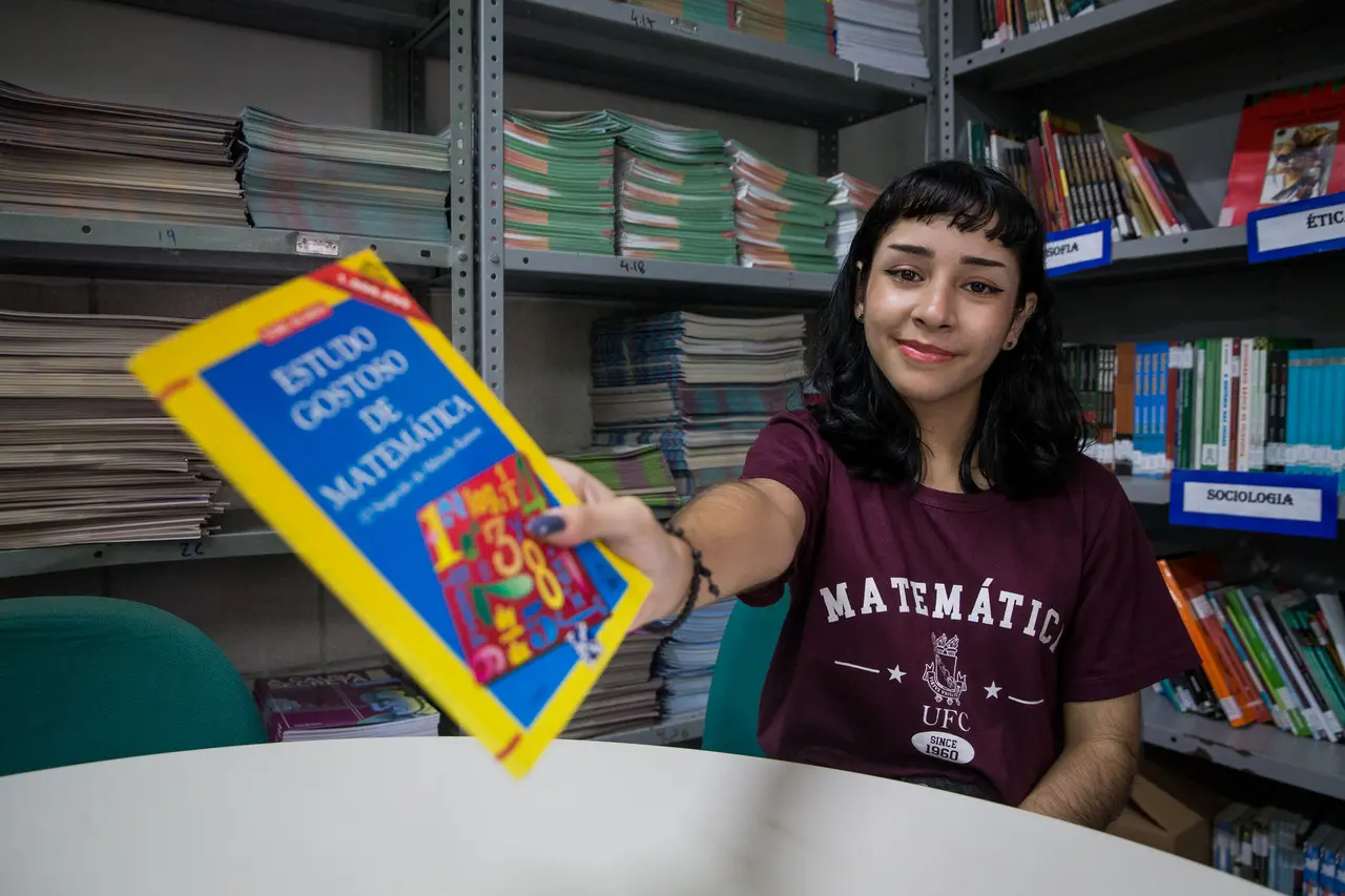 Estudante em biblioteca segura o livro “Estudo Gostoso de Matemática”, vestindo camisa com a palavra “Matemática” e o logo da UFC