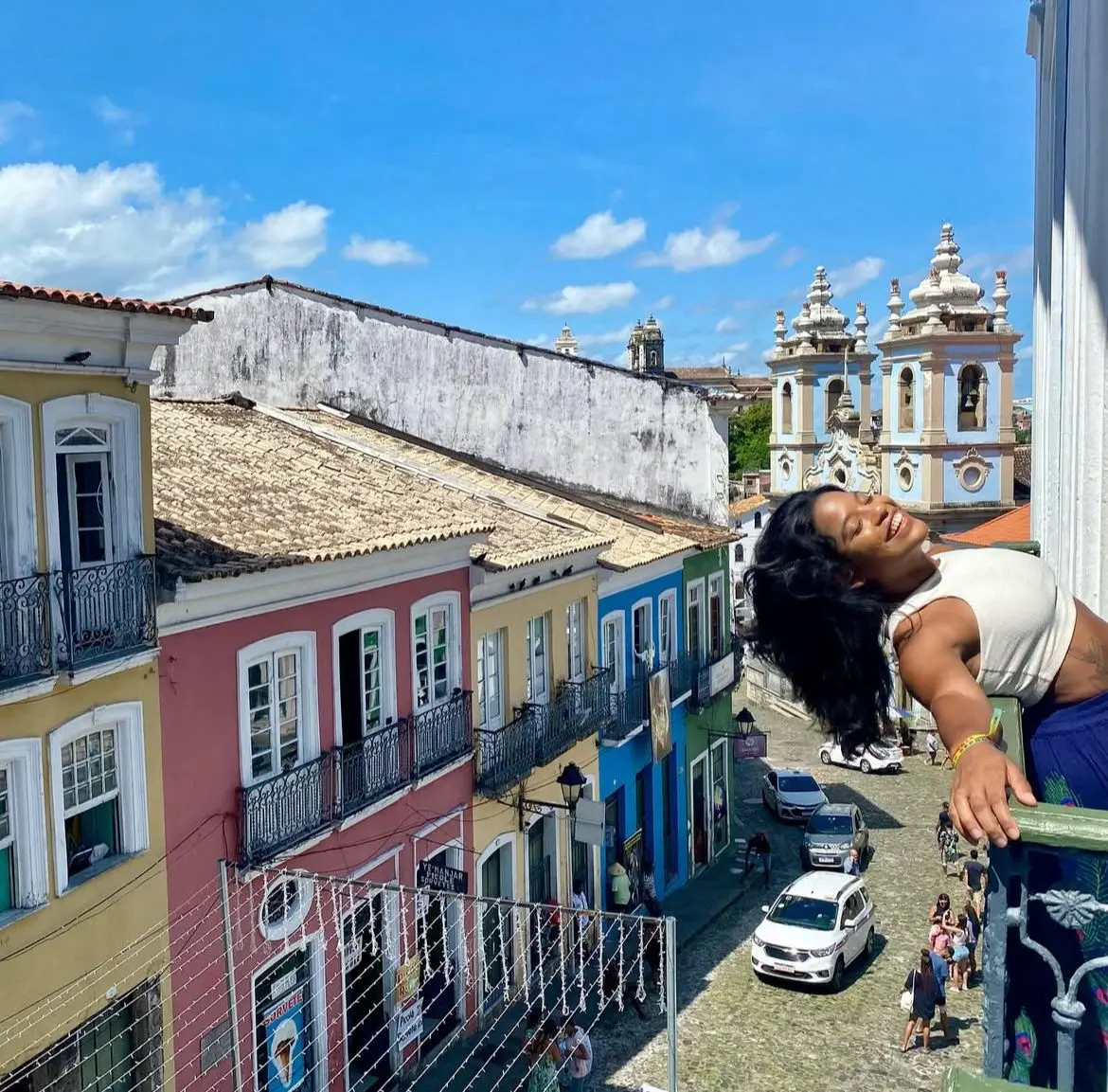 Juliana Marins sorrindo, uma mulher negra, inclinada para fora de uma sacada colorida no Pelourinho, em Salvador, com igrejas ao fundo e céu azul