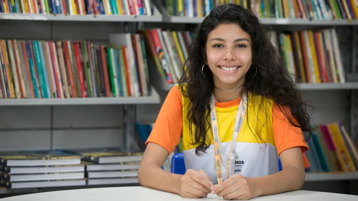 Jovem estudante sorri sentada à mesa em uma biblioteca, com estantes de livros ao fundo