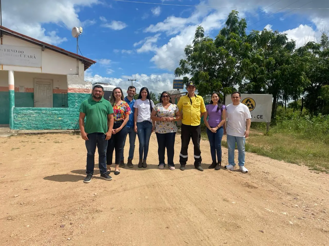 A imagem mostra um grupo de oito pessoas em pé em um terreno de terra batida, com um céu azul e nuvens brancas ao fundo. À esquerda, há um prédio de um andar com paredes brancas e uma base verde/azul, e uma placa parcialmente visível que diz 