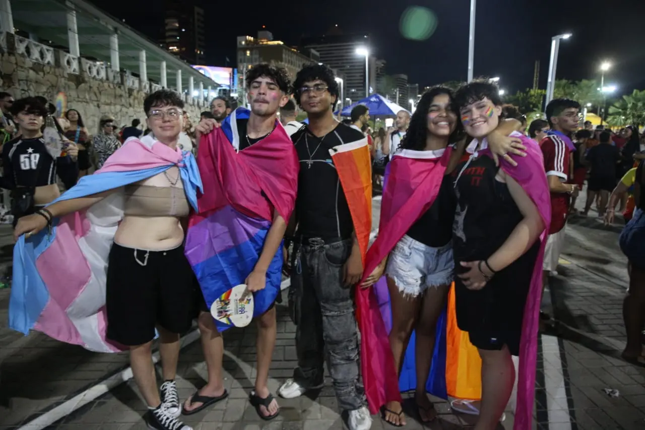 Grupo de cinco jovens posa sorridente para a foto em um evento noturno ao ar livre, com bandeiras coloridas representando diferentes identidades da comunidade LGBTQIA+ — incluindo as bandeiras trans, bissexual, lésbica e pansexual. Eles estão abraçados e usam as bandeiras como capas, demonstrando orgulho e união. Ao fundo, outras pessoas também participam da celebração, com prédios iluminados e postes de luz compondo a paisagem urbana. O clima é de festa, inclusão e diversidade.