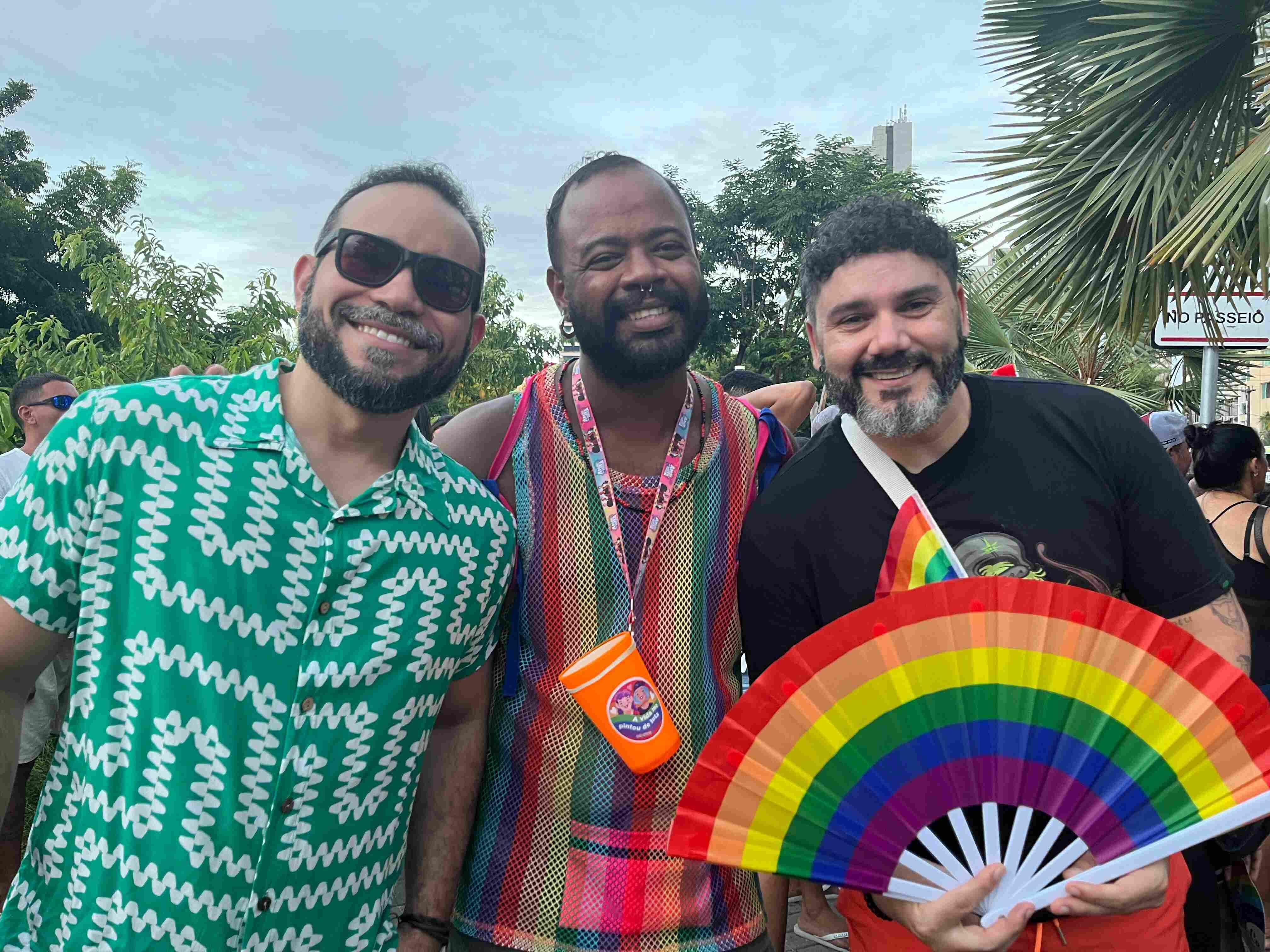 Três homens posam sorridentes em um evento ao ar livre durante o dia, cercados por outras pessoas e vegetação. O homem à esquerda veste uma camisa verde estampada e óculos escuros; o do centro usa uma regata colorida em tecido transparente e carrega um copo laranja pendurado no pescoço; o da direita segura um leque com as cores do arco-íris e tem uma pequena bandeira LGBTQIA+ presa à alça transversal de sua bolsa. O clima é festivo, com destaque para os elementos que celebram o orgulho e a diversidade sexual. Ao fundo, há árvores, palmeiras e uma placa de rua parcialmente visível.