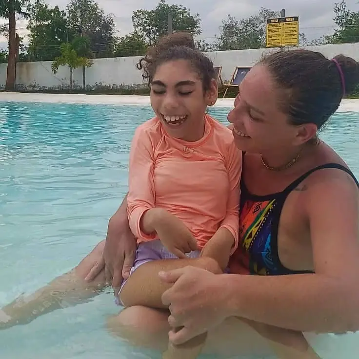 A fotografia mostra mãe e filha numa piscina. A criança está de laranja no colo da mãe, as duas estão sorrindo.
