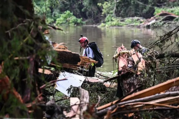 Dois socorristas equipados com capacetes, mochilas e câmeras presas à cabeça caminham por uma área devastada por enchente, repleta de galhos quebrados, troncos de árvores arrancados e destroços de madeira acumulados. Um dos profissionais está parcialmente submerso na água barrenta de um rio ao fundo, enquanto o outro avança por entre os escombros. O cenário é de floresta alagada, com vegetação densa e sinais claros da força da enchente que destruiu a paisagem natural.