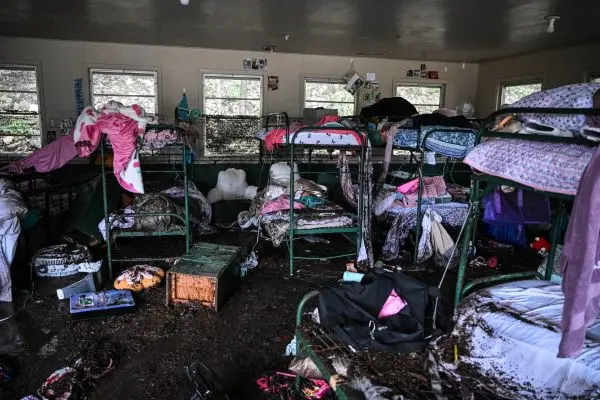 Imagem de um dormitório em estado de destruição, com vários beliches de metal revirados e cobertos por lama e objetos espalhados. As camas estão desorganizadas, com colchões sujos, roupas de cama e pertences pessoais molhados e jogados no chão. A terra e a lama cobrem o piso, indicando que o local foi afetado por uma enchente. As janelas mostram árvores do lado de fora, sugerindo que o local está em uma área arborizada. Fotografias e objetos pessoais ainda estão afixados nas paredes, contrastando com o cenário de devastação no interior do cômodo.