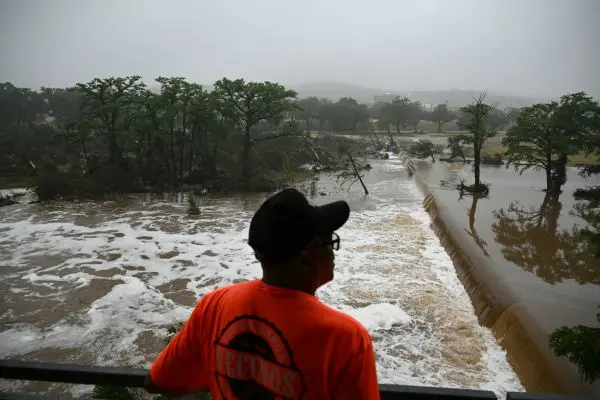 Homem com camiseta laranja e boné preto observa uma enchente a partir de uma estrutura elevada. À frente, uma forte correnteza transborda uma barragem, com grande volume de água barrenta e espumosa avançando sobre a vegetação. Árvores estão parcialmente submersas ou caídas, evidenciando a força da inundação. O céu está nublado e a visibilidade reduzida, indicando tempo chuvoso e clima severo. Ao fundo, é possível ver uma área urbana parcialmente encoberta pela névoa.