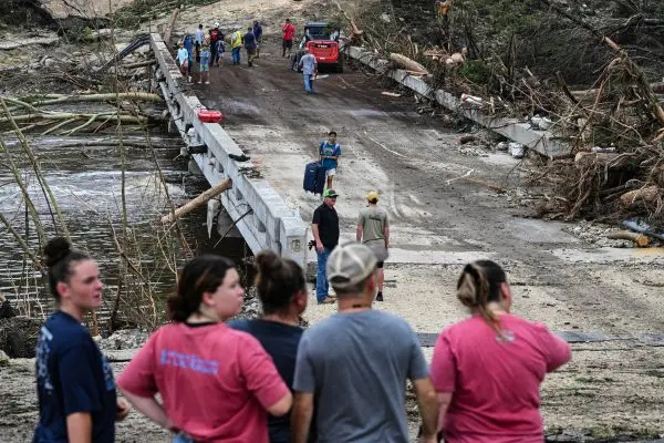 Grupo de pessoas observa uma ponte parcialmente destruída após uma enchente. A estrutura está coberta por lama e detritos, com troncos de árvores quebradas acumulados nas laterais. Algumas pessoas atravessam a ponte a pé, carregando pertences, enquanto outras permanecem na margem oposta. O cenário mostra sinais claros de devastação, com vegetação danificada e marcas da força da água. O ambiente é de reconstrução e avaliação dos danos, com moradores e trabalhadores presentes no local.