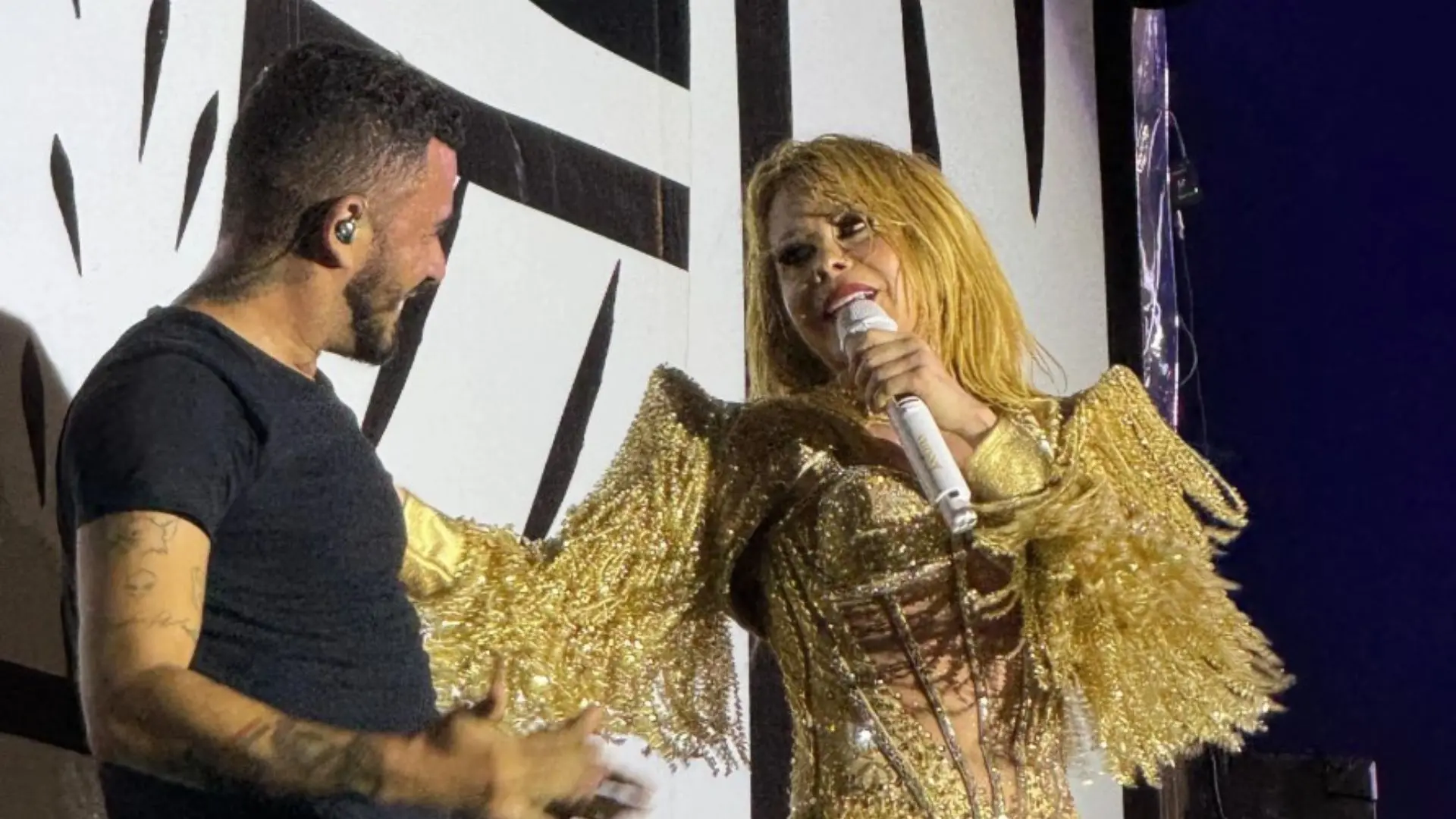 Apresentação de um Joelma e o intérprete Adriano Ruan no palco, com o público assistindo, destaque para a cantora com cabelo loiro e jaqueta dourada.