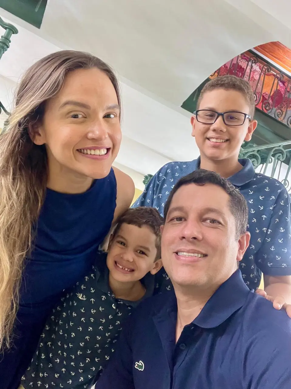 Uma selfie em close de uma família sorridente de quatro pessoas: um homem, uma mulher e dois meninos, posando em um ambiente interno com uma escada ao fundo. Todos estão olhando e sorrindo para a câmera.