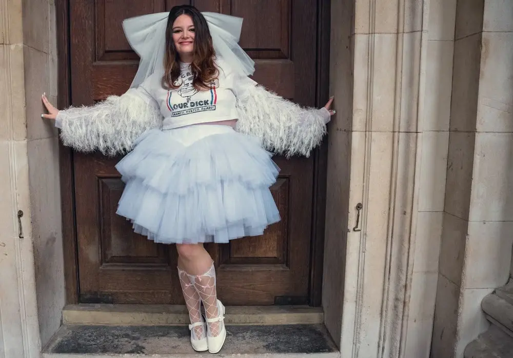 Atriz Megan Stalter caracterizada como Jessica posa para foto com saia de tule volumosa em frete a uma porta marrom