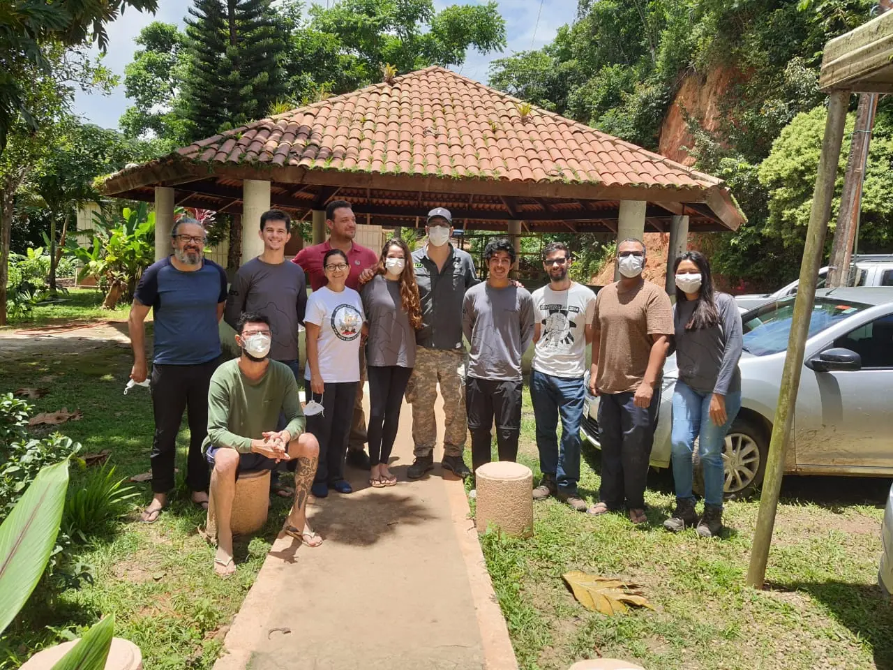 A foto mostra uma equipe de 12 pessoas, incluindo homens e mulheres. Quase todos usam máscaras. Eles estão em pé em um caminho pavimentado, com um telhado de telhas ao fundo. À direita, um carro prateado está estacionado, e à esquerda há árvores e folhagens. O ambiente parece ser um parque ou área verde.