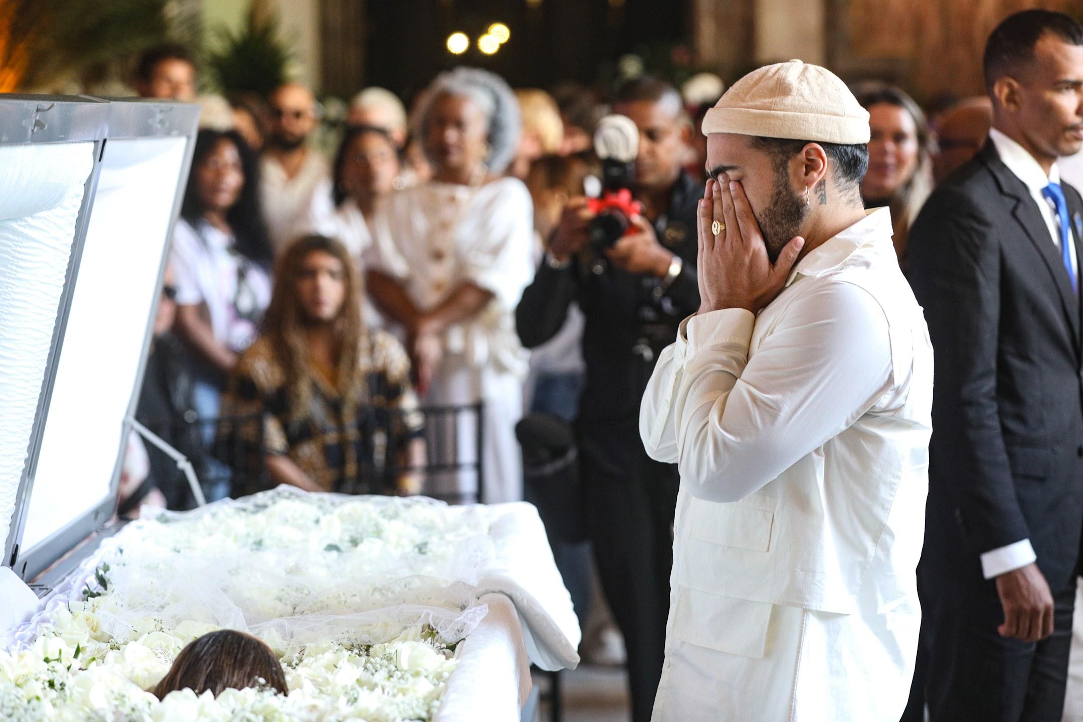Homem emocionado de cabelo e barba escuros, usando roupa branca e chapéu branco, presta homenagem em funeral com muitas pessoas ao fundo, em momento de luto e despedida.