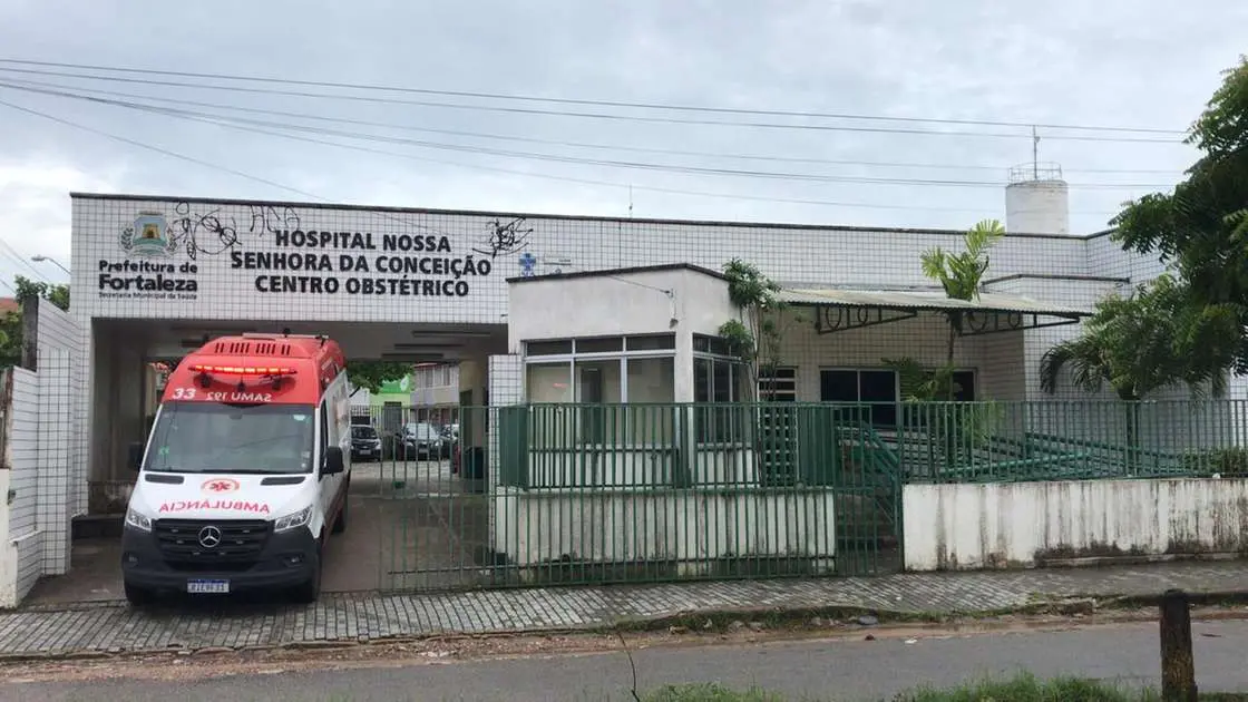A foto mostra a frente do Hospital Nossa Senhora da Conceição em Fortaleza. O prédio de um andar, branco, tem uma placa com o nome do hospital e o logo da Prefeitura de Fortaleza. Uma ambulância branca e vermelha está estacionada em frente ao prédio, atrás de uma cerca de metal. O céu está nublado.
