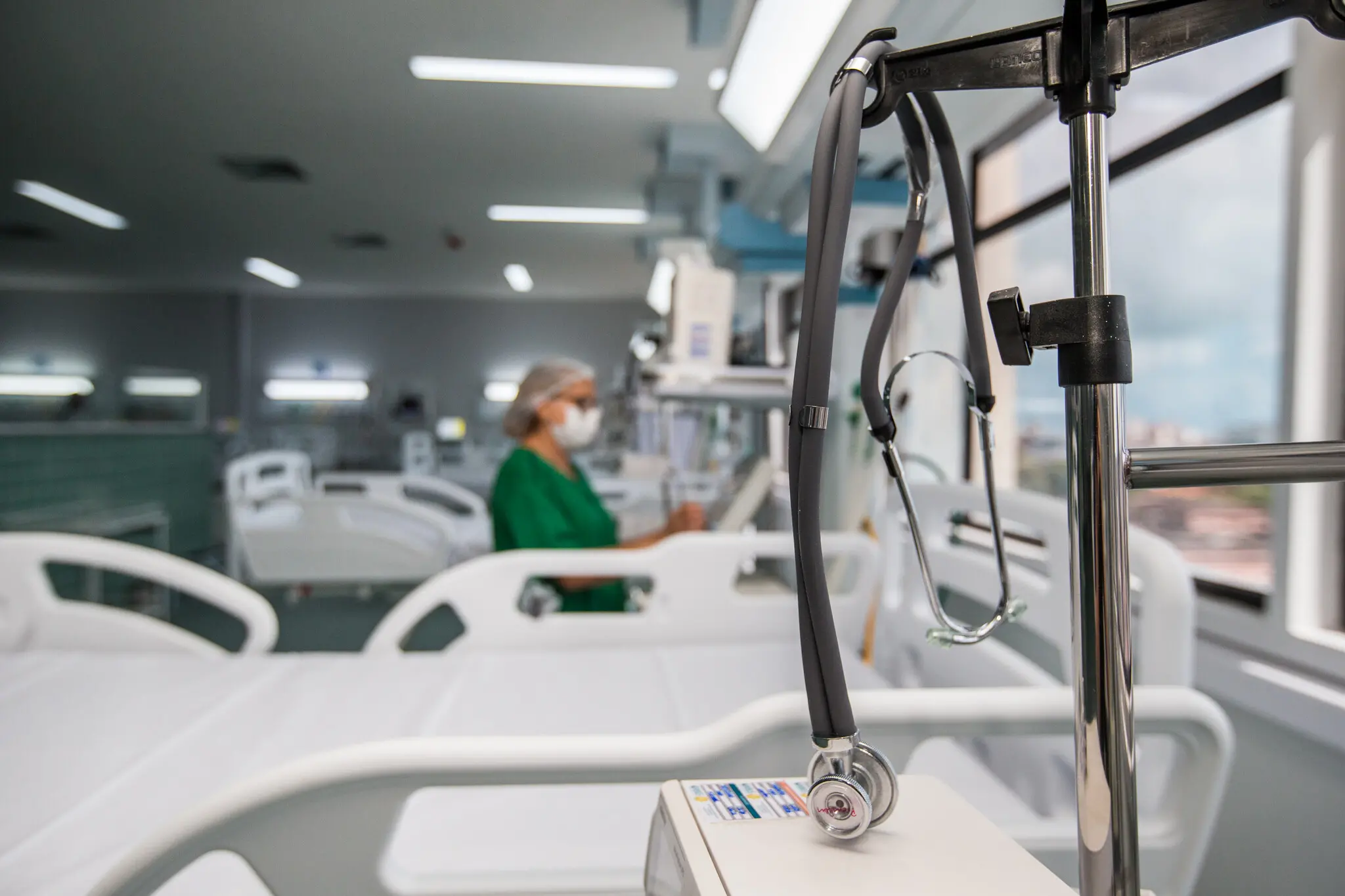 Novo leito de internação do IJF vazio, com estetoscópio ao lado, em foco, e profissional de saúde ao fundo em desfoque