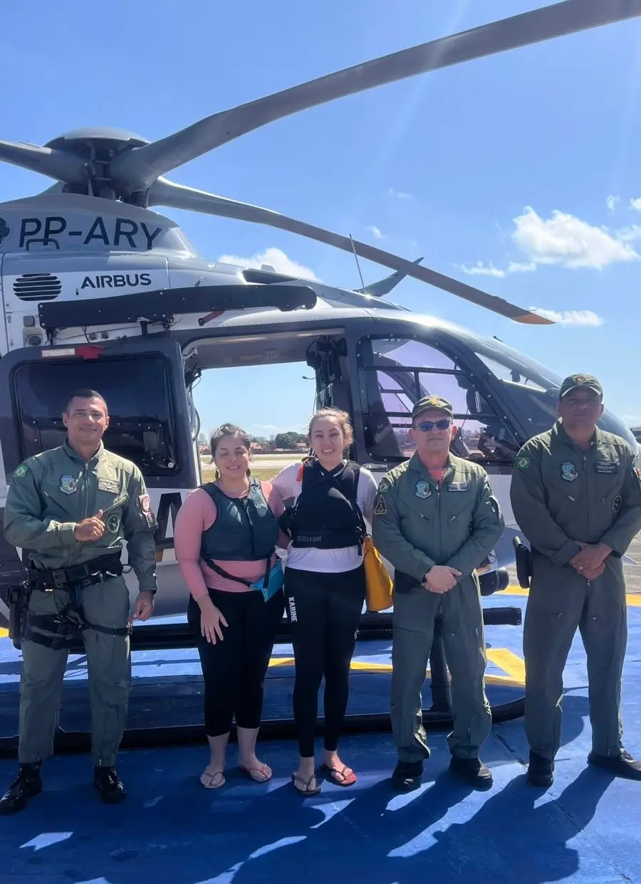 Foto de duas mulheres resgatadas e três profissionais da Ciopaer que atuaram no resgate, com um helicóptero ao fundo