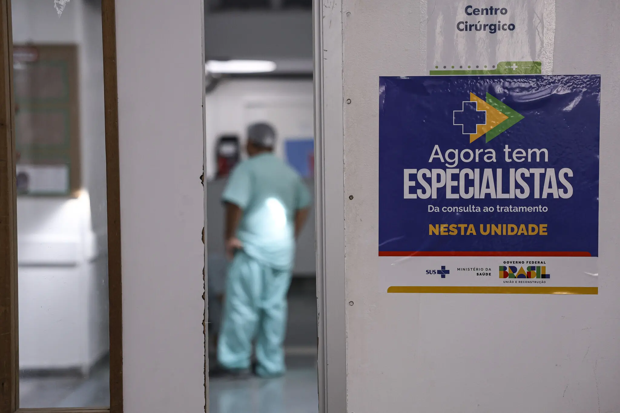 Porta de estabelecimento de saúde com placa do programa Agora Tem Especialistas, do Ministério da Saúde, e profissionais de saúde ao fundo, desfocados