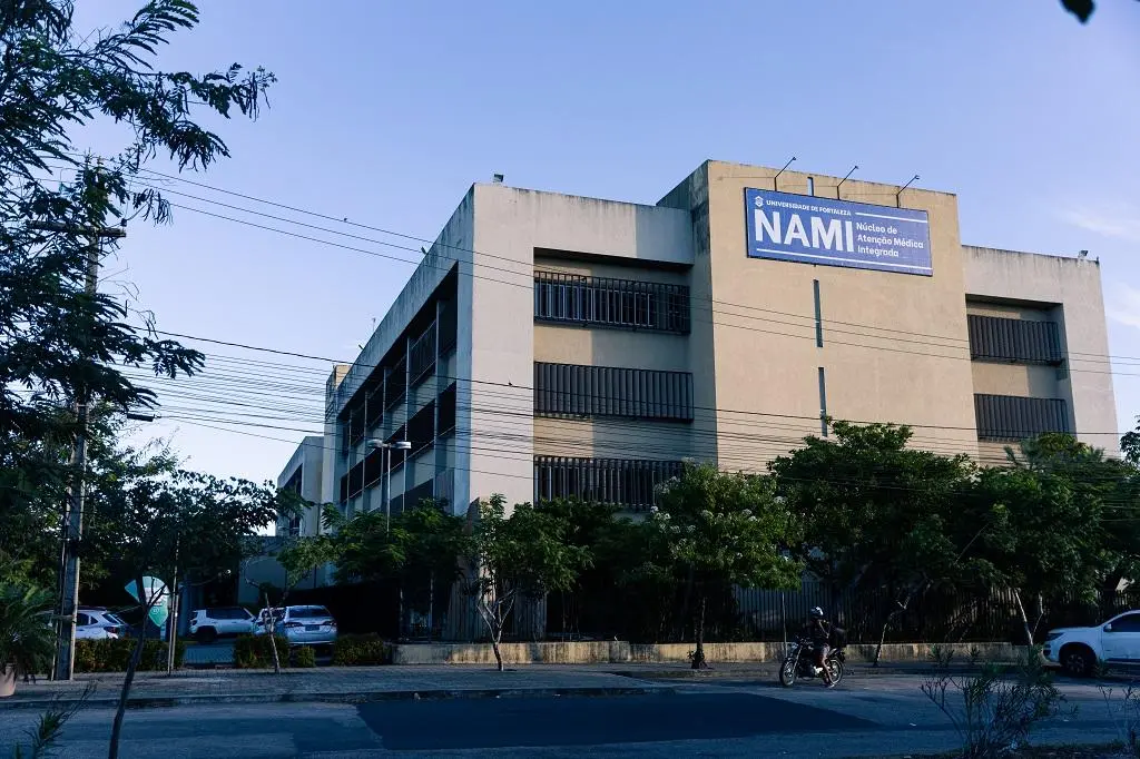 A foto mostra a fachada do prédio do NAMI (Núcleo de Atenção Médica Integrada) da Universidade de Fortaleza (UNIFOR). O prédio é de cor clara e possui janelas escuras com grades. Um grande banner azul com o nome 