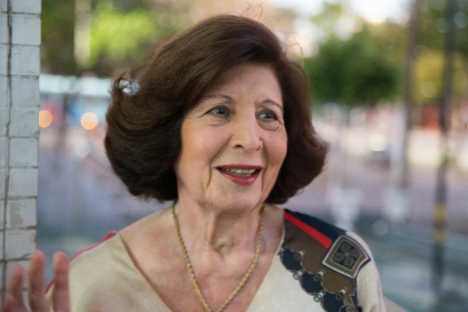 A foto mostra uma mulher de 80 anos, com cabelos castanhos escuros e médios. Ela tem olhos claros e usa um batom vermelho enquanto sorri levemente. A mulher veste uma blusa clara com detalhes verdes, marrons e vermelhos no lado esquerdo do ombro.