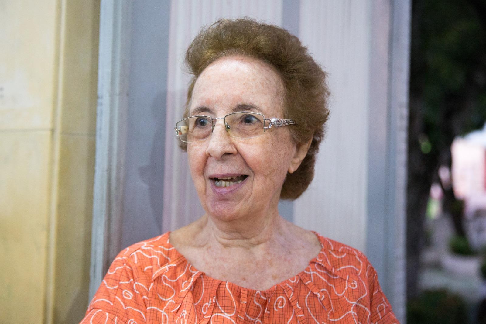 A foto mostra uma mulher de 81 anos, com cabelos curtos e loiro escuro. Ela usa um óculos com detalhes prateados. Ela sorri levemente.