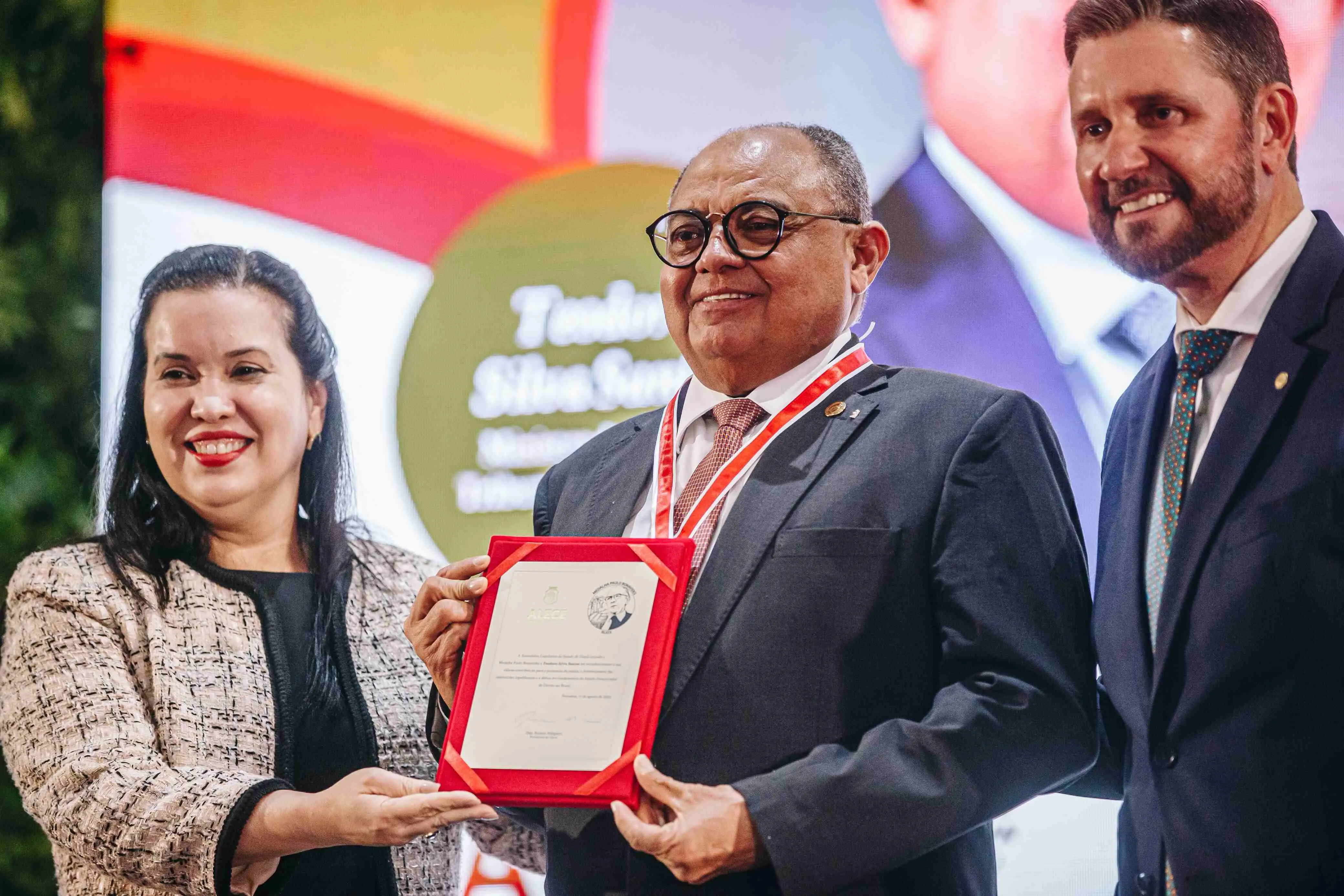 Imagem mostra um homem de óculos, pele morena e cabelos curtos grisalhos, vestindo terno escuro, camisa branca e gravata vermelha com padrão geométrico. Ele está em frente a um painel da Assembleia Legislativa do Ceará (ALECE) com o logotipo da “Medalha Paulo Bonavides” ao fundo, posando para foto em ambiente de evento formal.