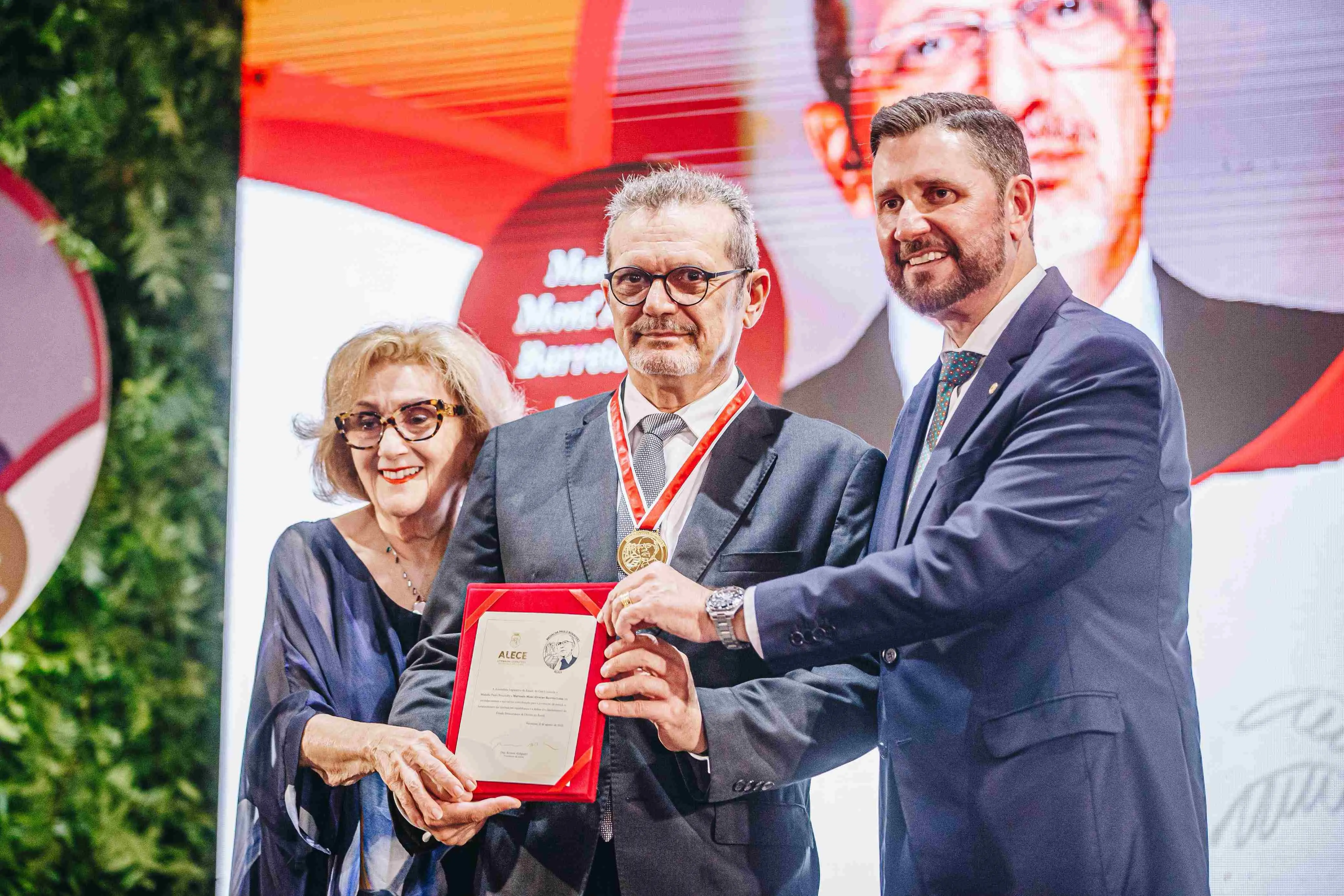Imagem mostra um homem de óculos arredondados, cabelo curto grisalho e barba rala, vestindo terno escuro, camisa clara e gravata azul, concedendo entrevista para a imprensa. Ele está cercado por vários microfones e gravadores. Ao fundo, há um painel da Assembleia Legislativa do Ceará (ALECE) e iluminação em tom avermelhado.