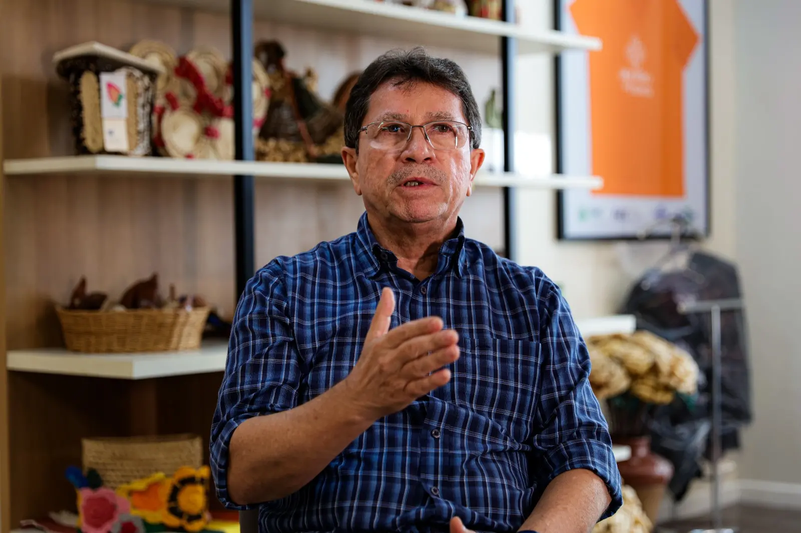 Imagem mostra Alci, um homem de meia-idade, usando óculos e camisa xadrez azul, sentado em ambiente interno, gesticulando com a mão direita. Ao fundo, há prateleiras com cestos de palha e artesanatos, além de um quadro com camiseta laranja emoldurada. Este é o seu escritório no Sebrae/CE.