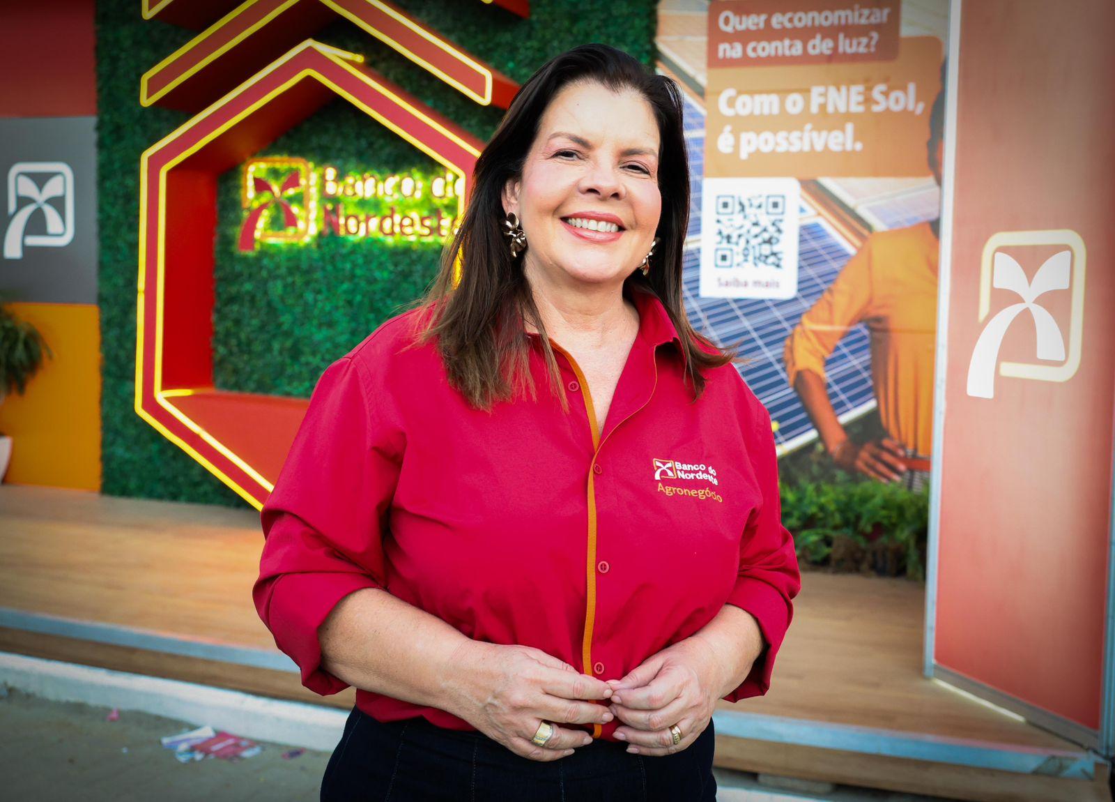 Mulher de cabelos castanhos e lisos, sorrindo, veste camisa social vermelha com o logotipo do Banco do Nordeste e a palavra “Agronegócio” bordada. Ao fundo, há um estande com elementos visuais do banco, incluindo logotipo iluminado, painel verde com vegetação artificial e cartaz sobre energia solar.