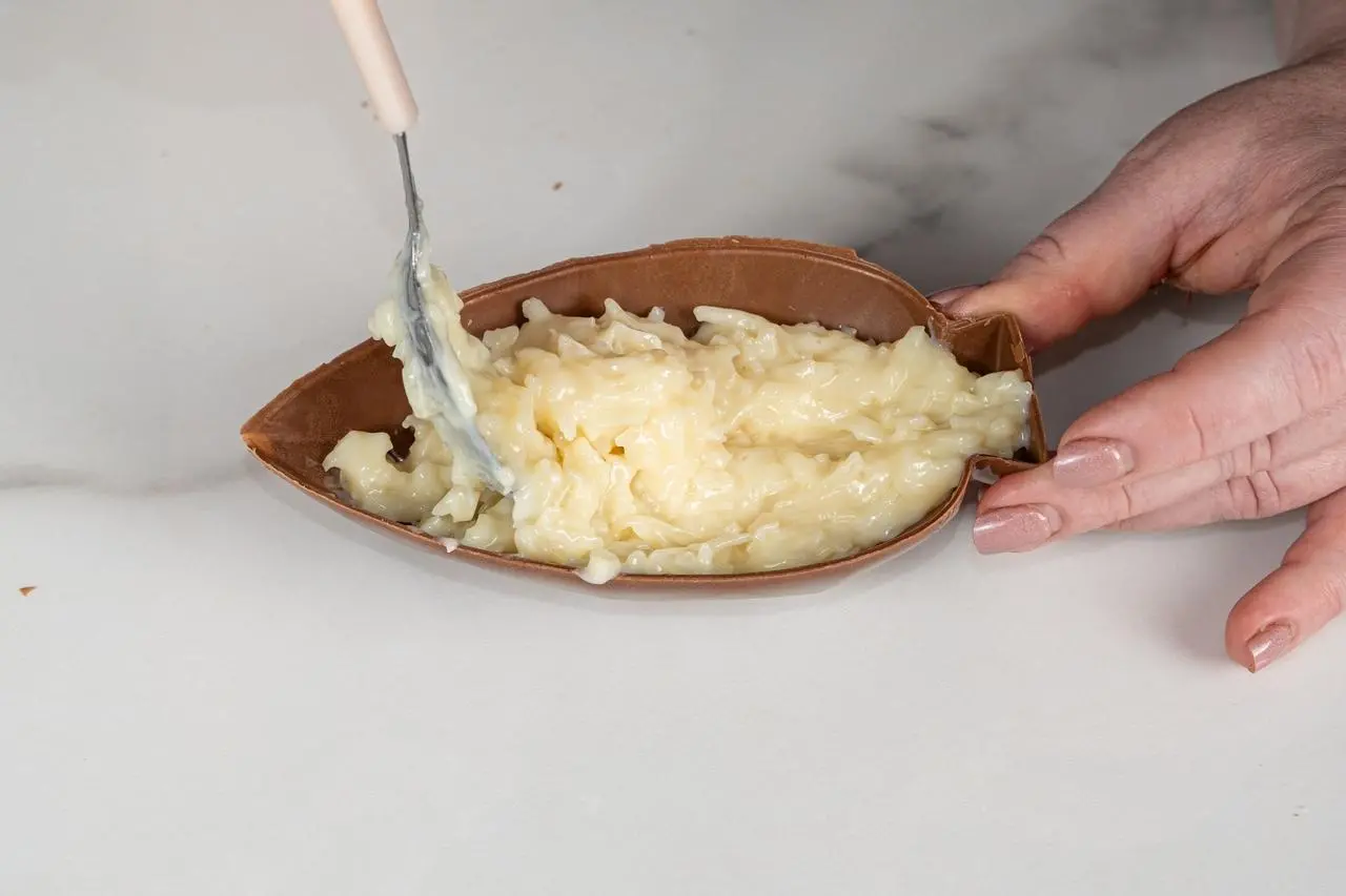 Imagem mostra mãos femininas recheando uma casca de ovo de chocolate artesanal com cocada caseira cremosa branca, utilizando uma colher em superfície de mármore clara.