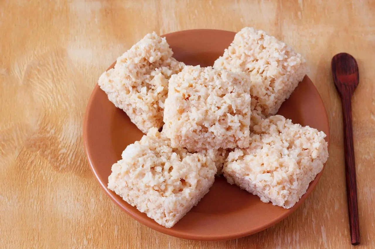 Cinco pedaços quadrados de cocada caseira de corte dispostos sobre um prato marrom, com uma pequena colher de madeira ao lado, em fundo de madeira clara