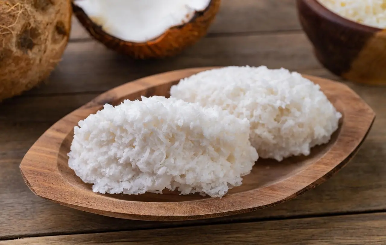 Imagem mostra close-up de duas cocadas caseiras brancas fofas e úmidas servidas em um pequeno prato de madeira clara, com metades de coco e coco ralado desfocado ao fundo.