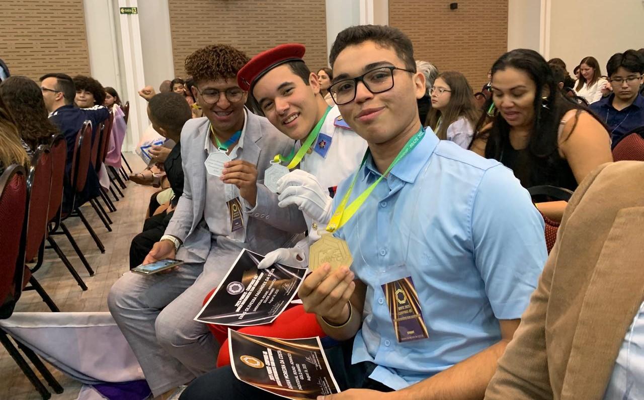 Três jovens sorrindo e segurando medalhas de ouro e prata. O da esquerda, com cabelo encaracolado e óculos, veste um terno cinza e segura uma medalha de prata. O do meio, com um chapéu vermelho, veste um uniforme formal com luvas brancas e mostra uma medalha de prata. O da direita, também de óculos, veste uma camisa polo azul e exibe uma medalha de ouro. Eles estão sentados em um auditório, em meio a uma plateia, e parecem estar em uma cerimônia de premiação.