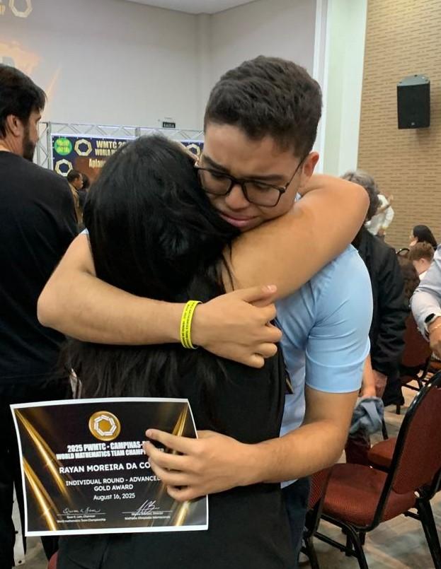 Um jovem de óculos e camisa polo azul está abraçando uma mulher com cabelos escuros, que está de costas para a câmera. Eles estão em um evento, com outras pessoas ao redor, e banners ao fundo indicam que se trata de uma competição de matemática.