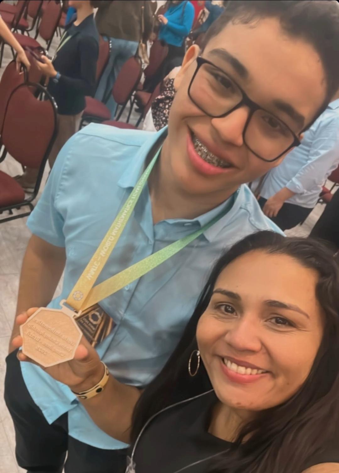 Um selfie tirado por uma mulher sorridente com cabelos castanhos escuros e brincos de argola, que segura uma grande medalha de bronze. Ao lado dela, um jovem de óculos e aparelho nos dentes, vestindo uma camisa polo azul, sorri para a câmera. Ele usa a mesma fita de pescoço, indicando que também participou do evento.