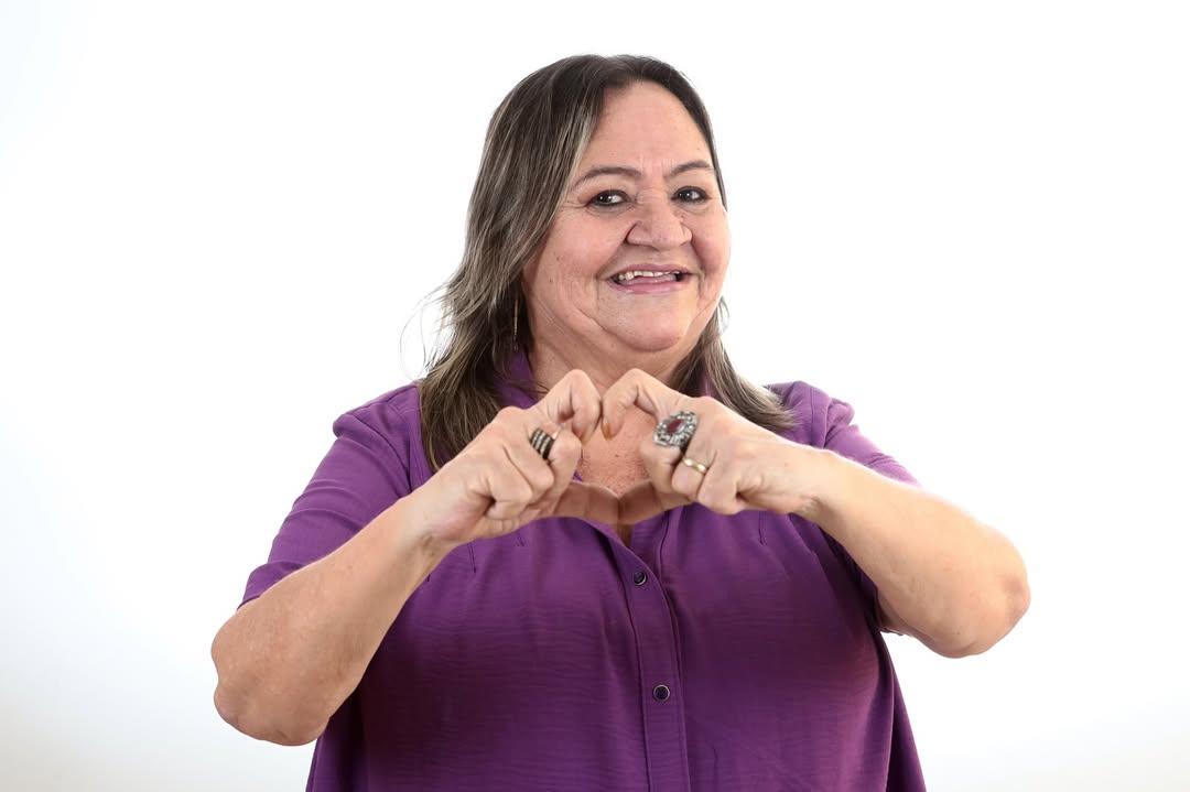 foto de janne ruth, bailarina e coreógrafa. ela veste roupa roxa, sorri e faz um coração com as mãos. a foto tem fundo branco.