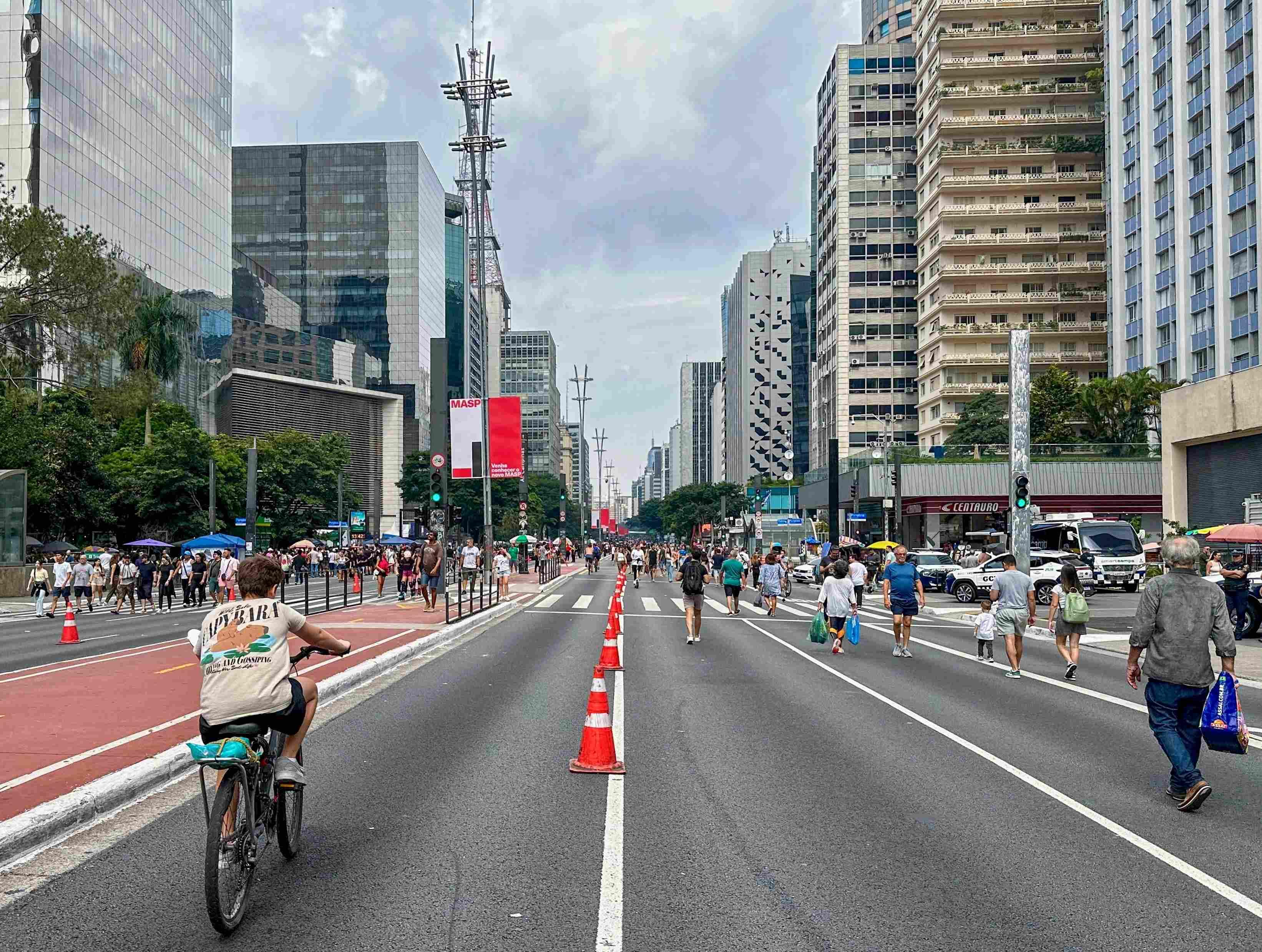 Imagem de uma rua urbana movimentada com pessoas caminhando e andando de bicicleta. A via é ladeada por prédios altos e possui cones de trânsito no centro, separando as faixas. Um ciclista com camisa bege aparece à esquerda, enquanto pedestres circulam pelos dois lados da rua. O céu está nublado e há árvores e vegetação ao longo das calçadas