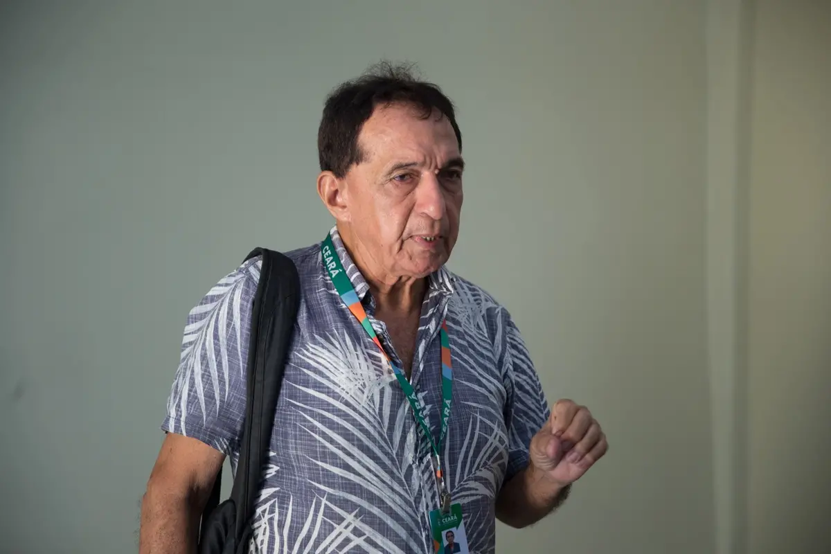 Homem usa camisa estampada em tons de cinza e branco, crachá no pescoço e mochila preta no ombro. Ele está em pé, diante de uma parede lisa, gesticulando com a mão direita.