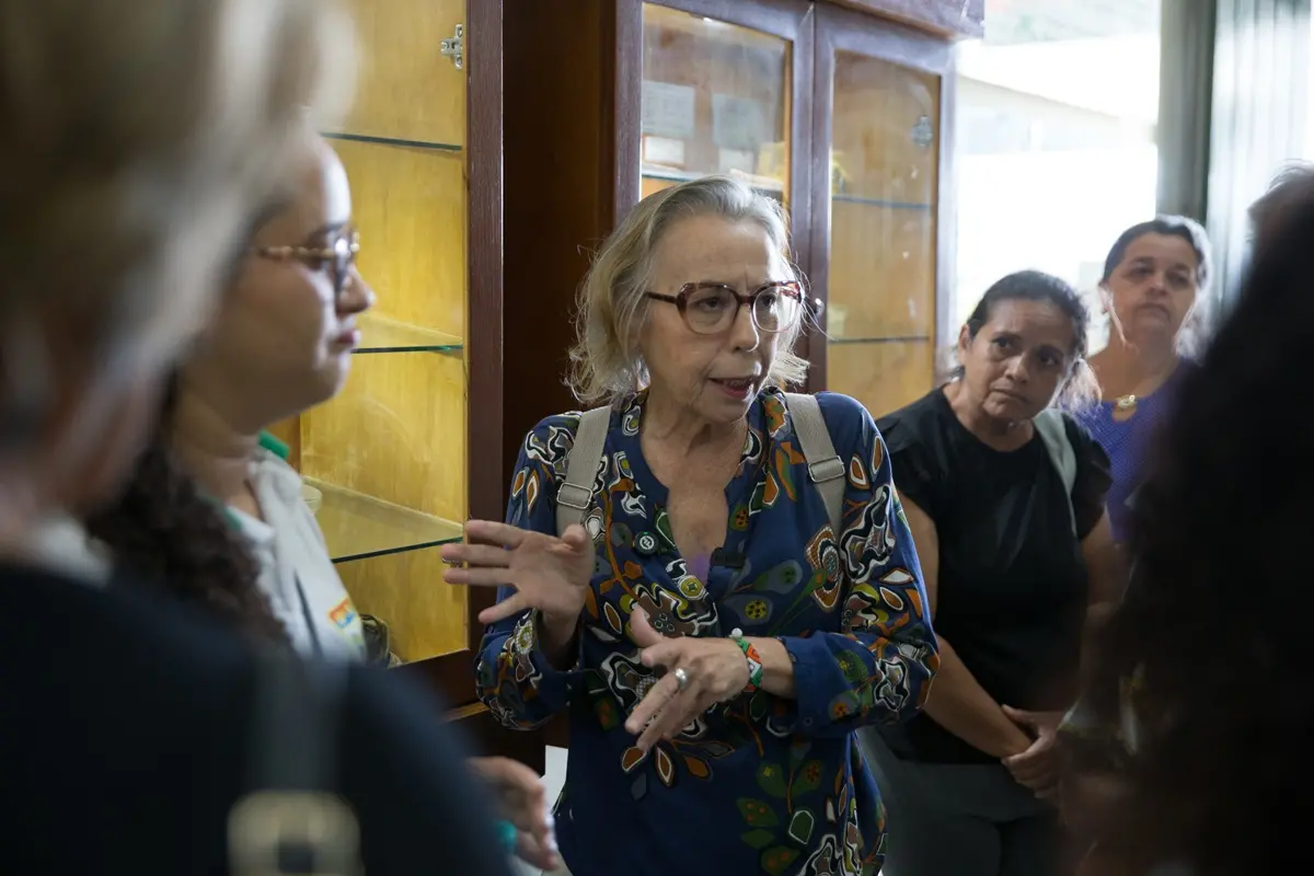 Mulher idosa, de cabelos claros e óculos de armação grossa, fala gesticulando com as mãos enquanto conversa com um grupo de pessoas em um ambiente interno, diante de uma estante de vidro.
