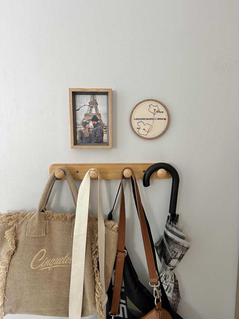 Cabideiro com roupas, guarda-chuva e bolsas penduradas. Pedurado na parece também estão um porta-retrato de família em Paris e um bordado à mão exposto em dispositivo circular