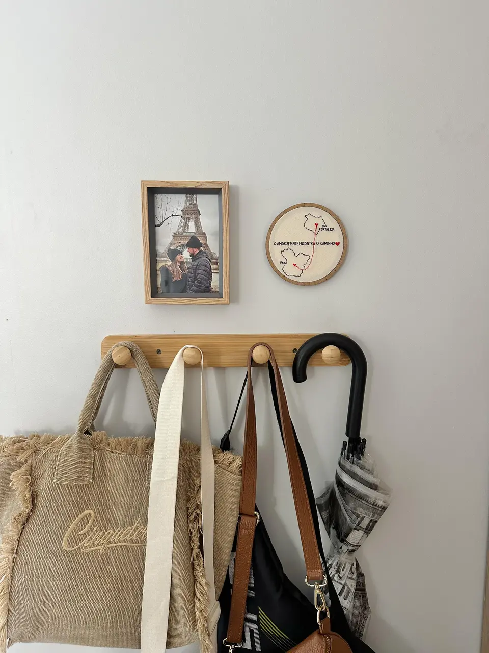 Cabideiro com roupas, guarda-chuva e bolsas penduradas. Pedurado na parece também estão um porta-retrato de família em Paris e um bordado à mão exposto em dispositivo circular
