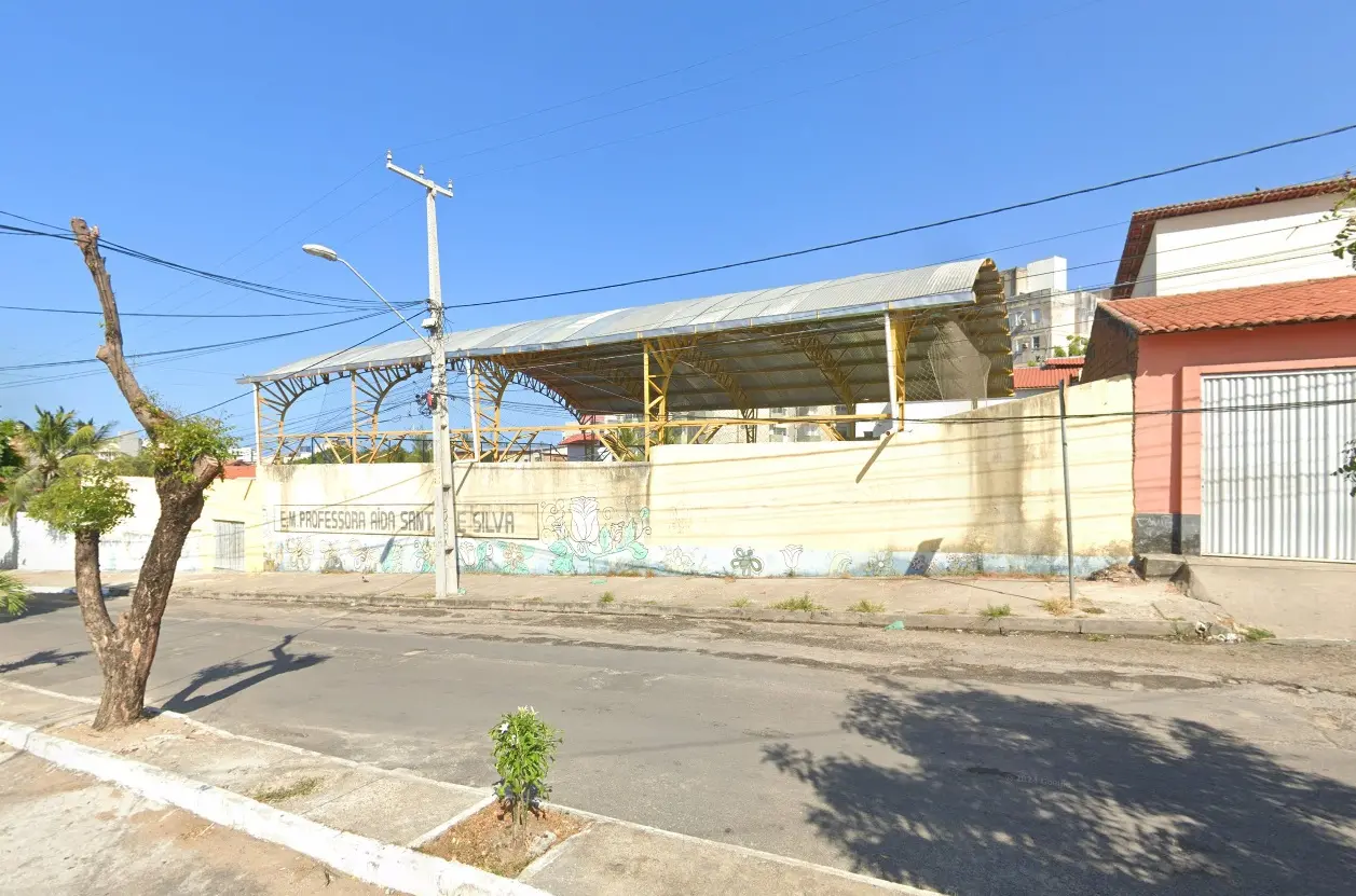 Vista externa de uma escola municipal, com muro pintado em tons claros e desenhos florais. Sobre o muro, há a identificação “E.M. Professora Aída Santos e Silva”. Ao fundo, vê-se uma quadra esportiva coberta com estrutura metálica amarela. A rua em frente está vazia, com árvores e pequenos arbustos na calçada.